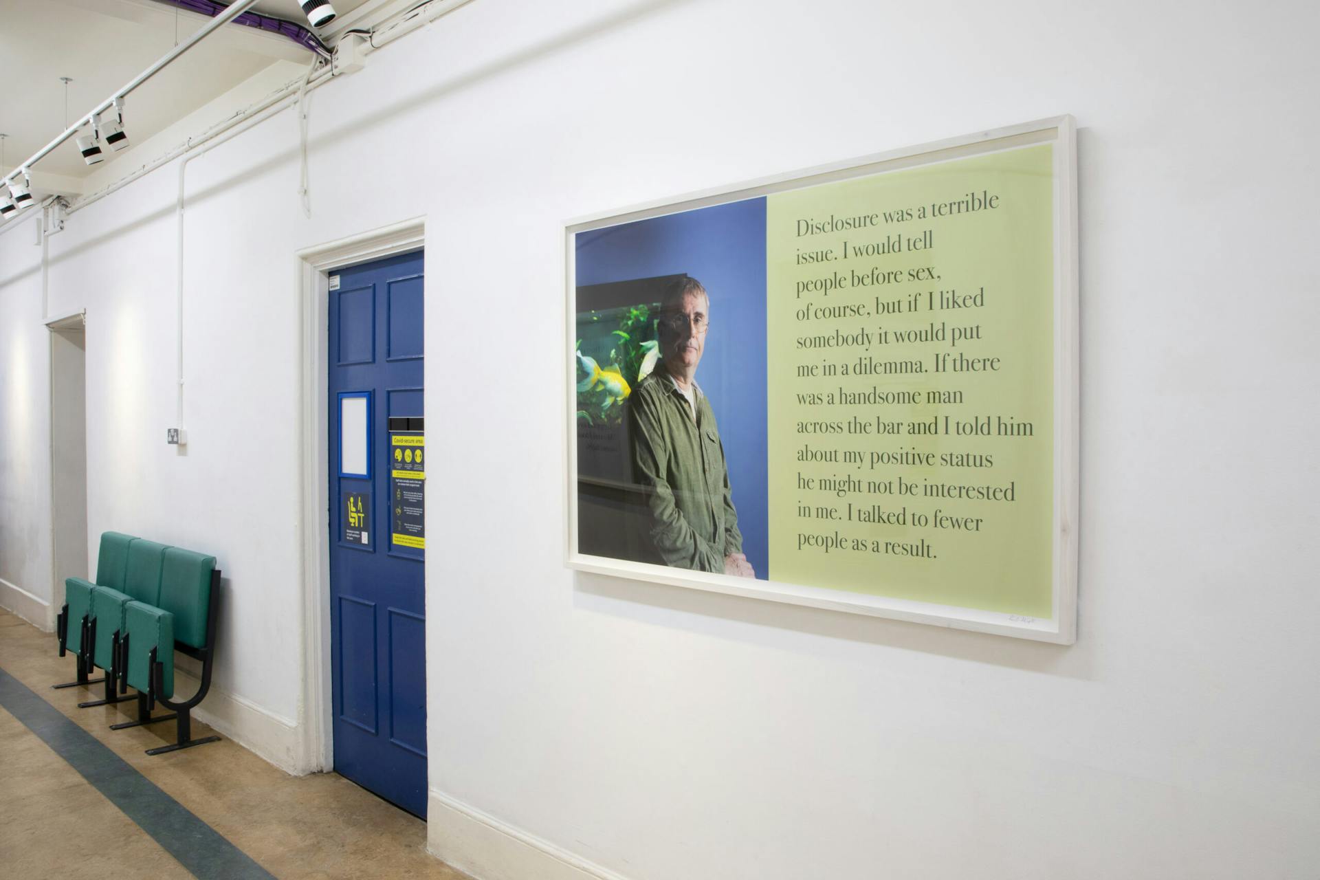 A hallway with white walls that display a framed photograph and text hang on the wall. The image features a man and the text discusses disclosure of his positive status and its impact on forming relationships. Green chairs are placed nearby.