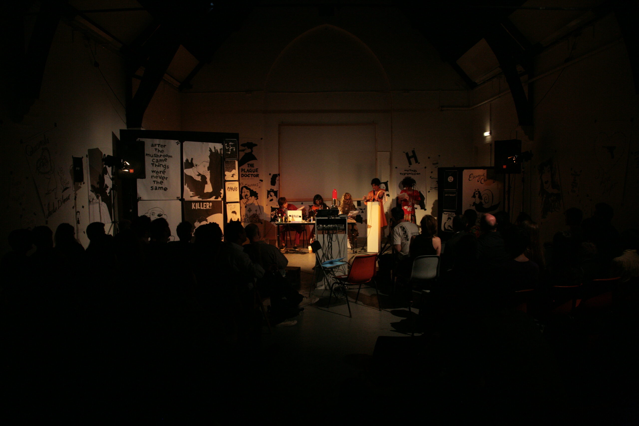 A wide angle view of a bank of performers wearing wigs and outlandish costumes, as seen through an audience.