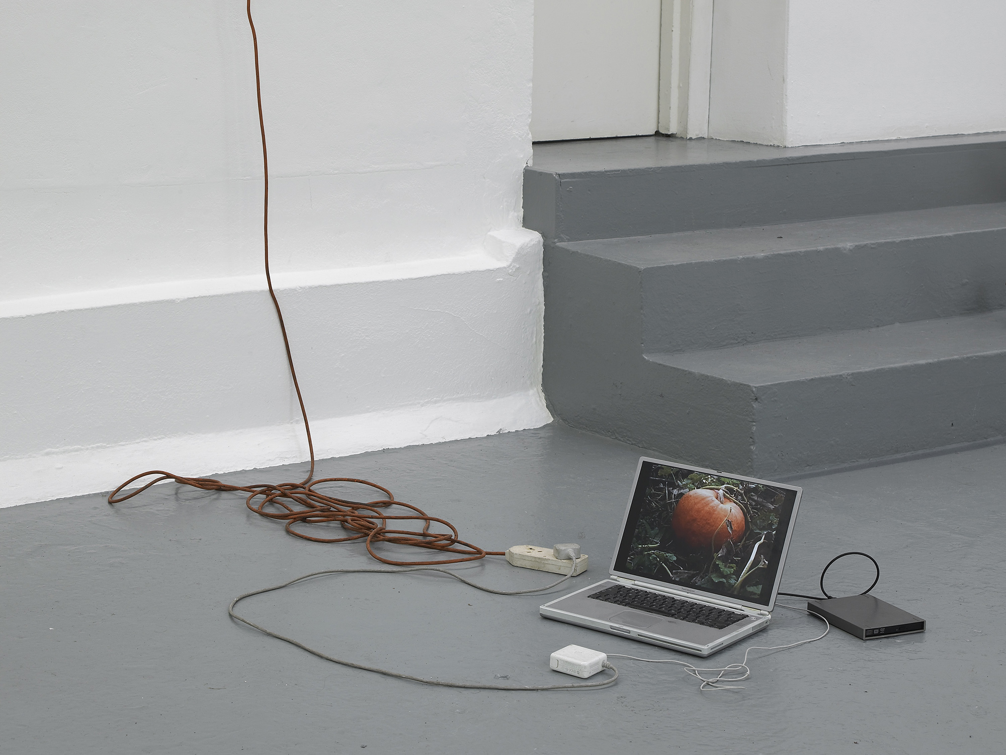 A laptop displaying an image of a fruit sits on a gray floor near steps, connected to a power adapter and external device, with a long brown extension cord trailing up the white wall.