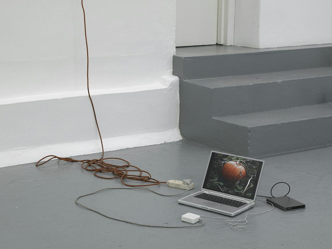 A laptop displaying an image of a fruit sits on a gray floor near steps, connected to a power adapter and external device, with a long brown extension cord trailing up the white wall.