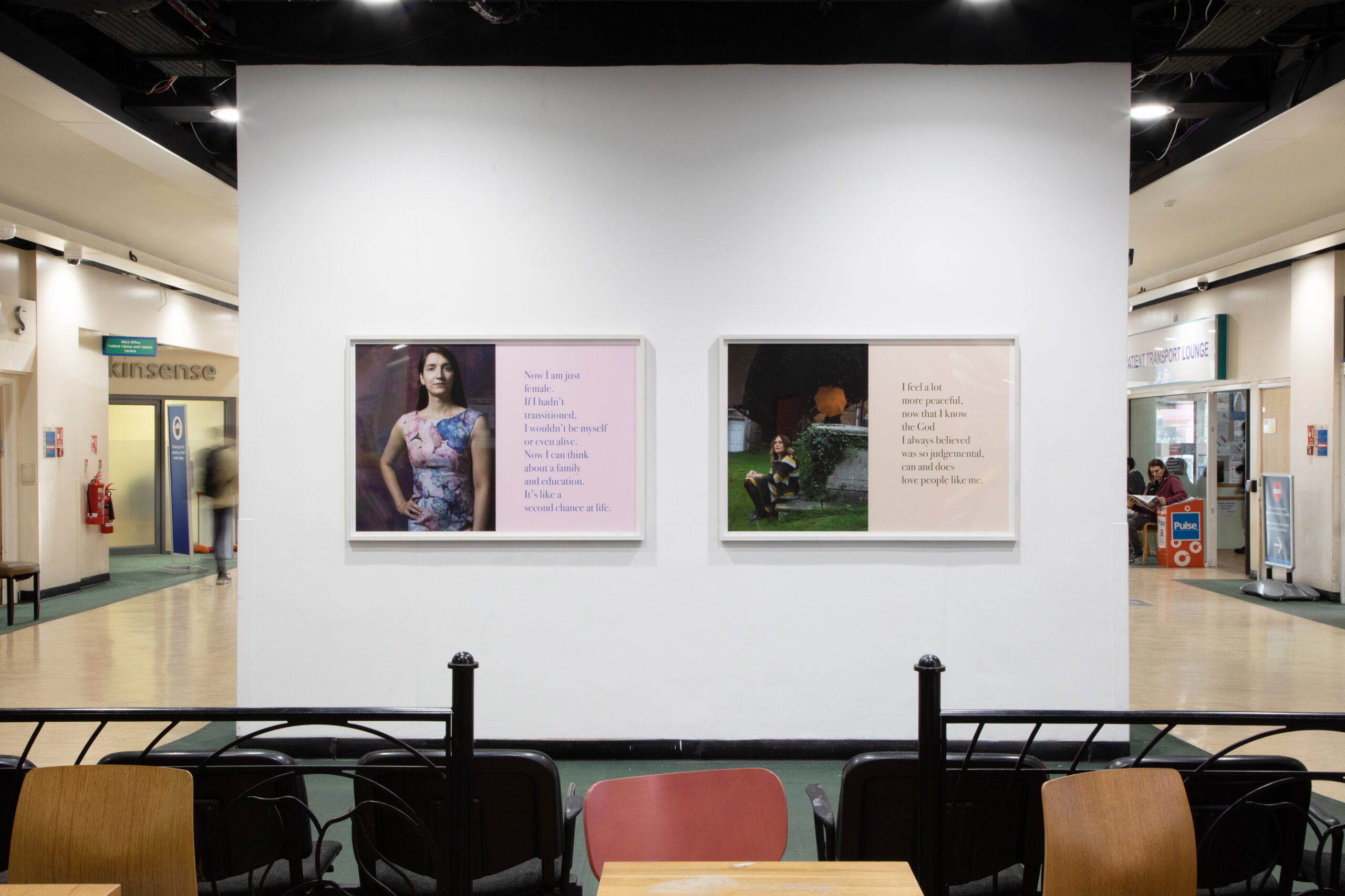 An exhibit featuring two framed pieces of art on a white wall within a hospital. The left artwork depicts a person in front of an illustrated background with text beside them. The right artwork displays an illustrated scene with text. People are visible in the background.