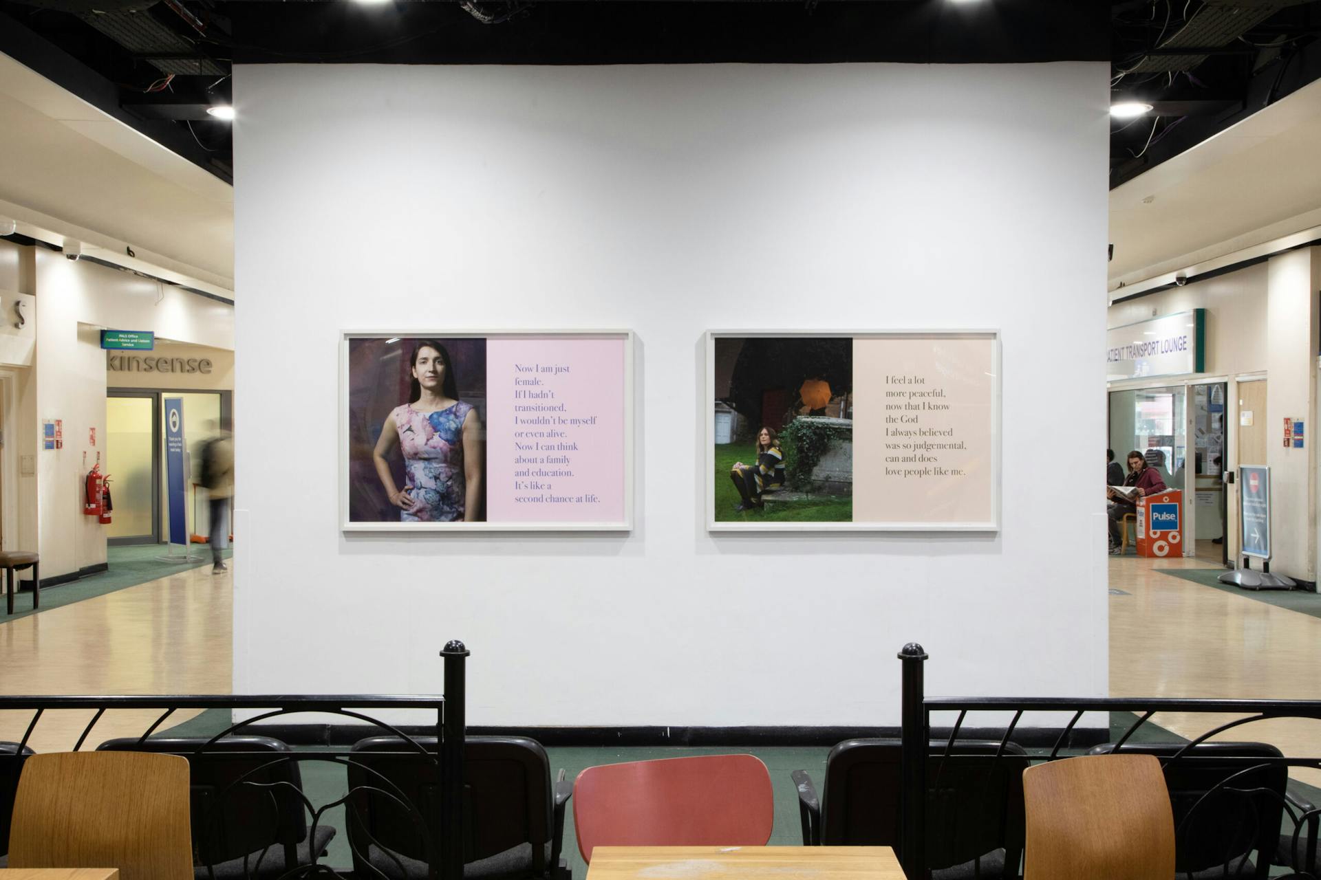 An exhibit featuring two framed pieces of art on a white wall within a hospital. The left artwork depicts a person in front of an illustrated background with text beside them. The right artwork displays an illustrated scene with text. People are visible in the background.
