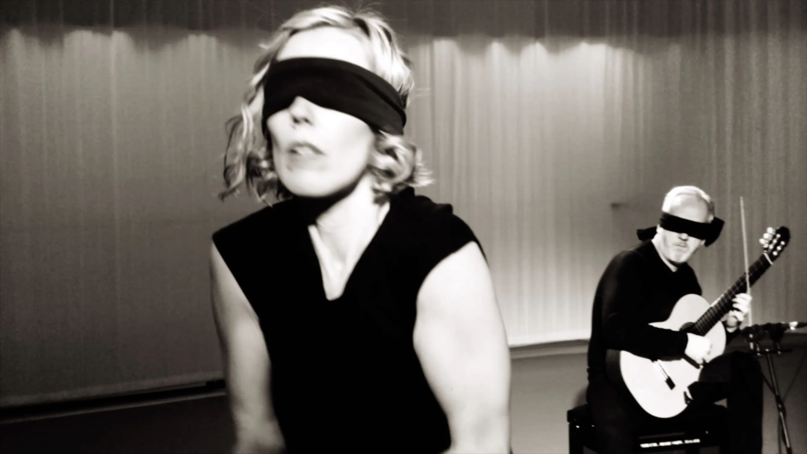 A black and white image of a woman with short hair, blindfolded, wearing a sleeveless top, moving energetically. In the background, a person sits playing an acoustic guitar. The setting appears to be a performance space.