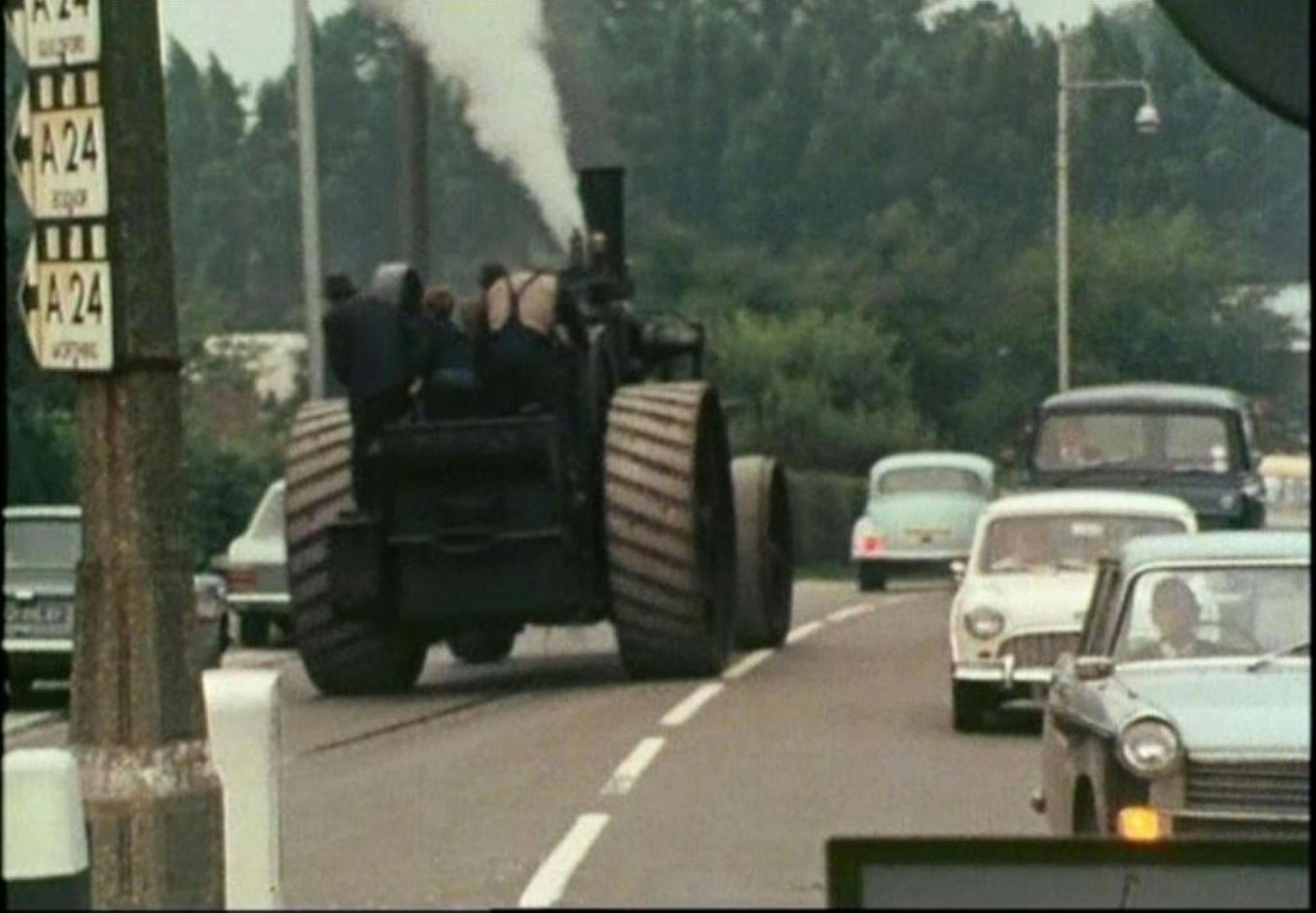 A steam-powered vehicle travels down a busy road, emitting white smoke from its chimney. Several people are riding it. Surrounding the vehicle are 1960s-era cars driving in both directions. A tall road sign is visible on the left. Trees line the road.