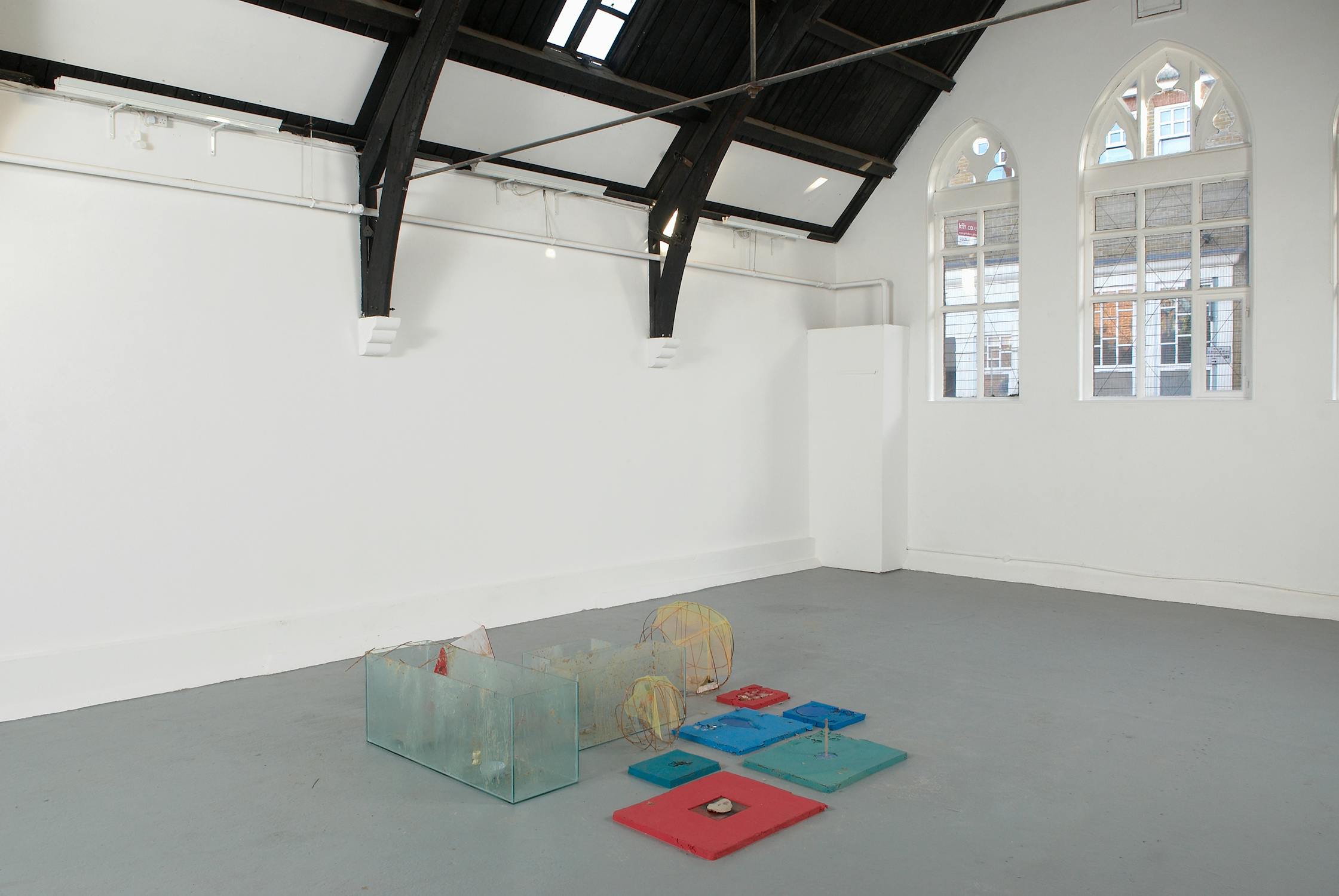 A wide angle photo of Studio Voltaire's arched gallery space. In the middle of the frame, a collection of three glass tanks smeared with porridge sit amongst a collection of low to the ground red, green and blue cast silicon blocks. amongst these objects sit two open-weave twine structures topped with scraps of yellow net