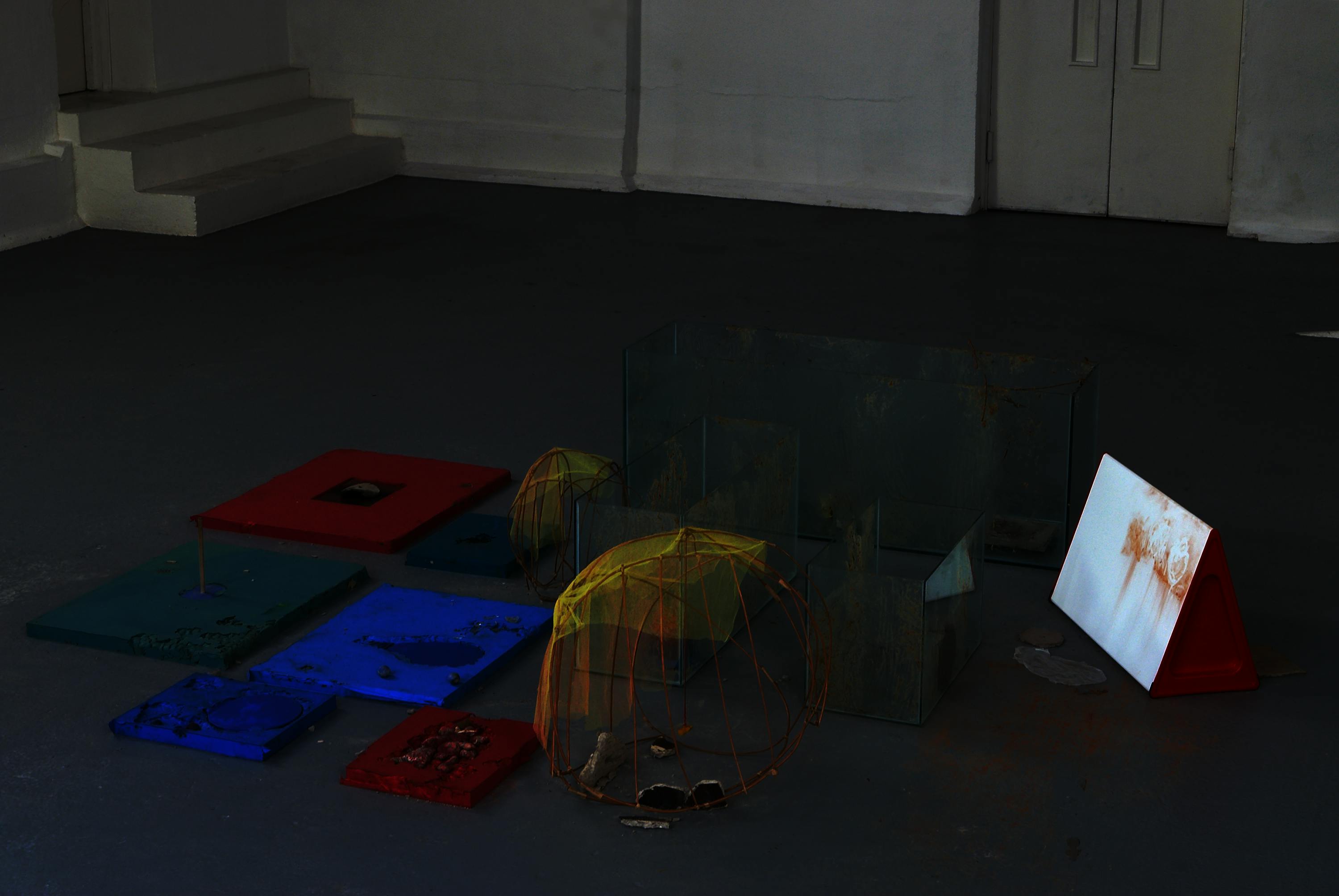 three glass tanks smeared with porridge sit amongst a collection of low to the ground red, green and blue cast silicon blocks. amongst these objects sit two open-weave twine structures topped with scraps of yellow net