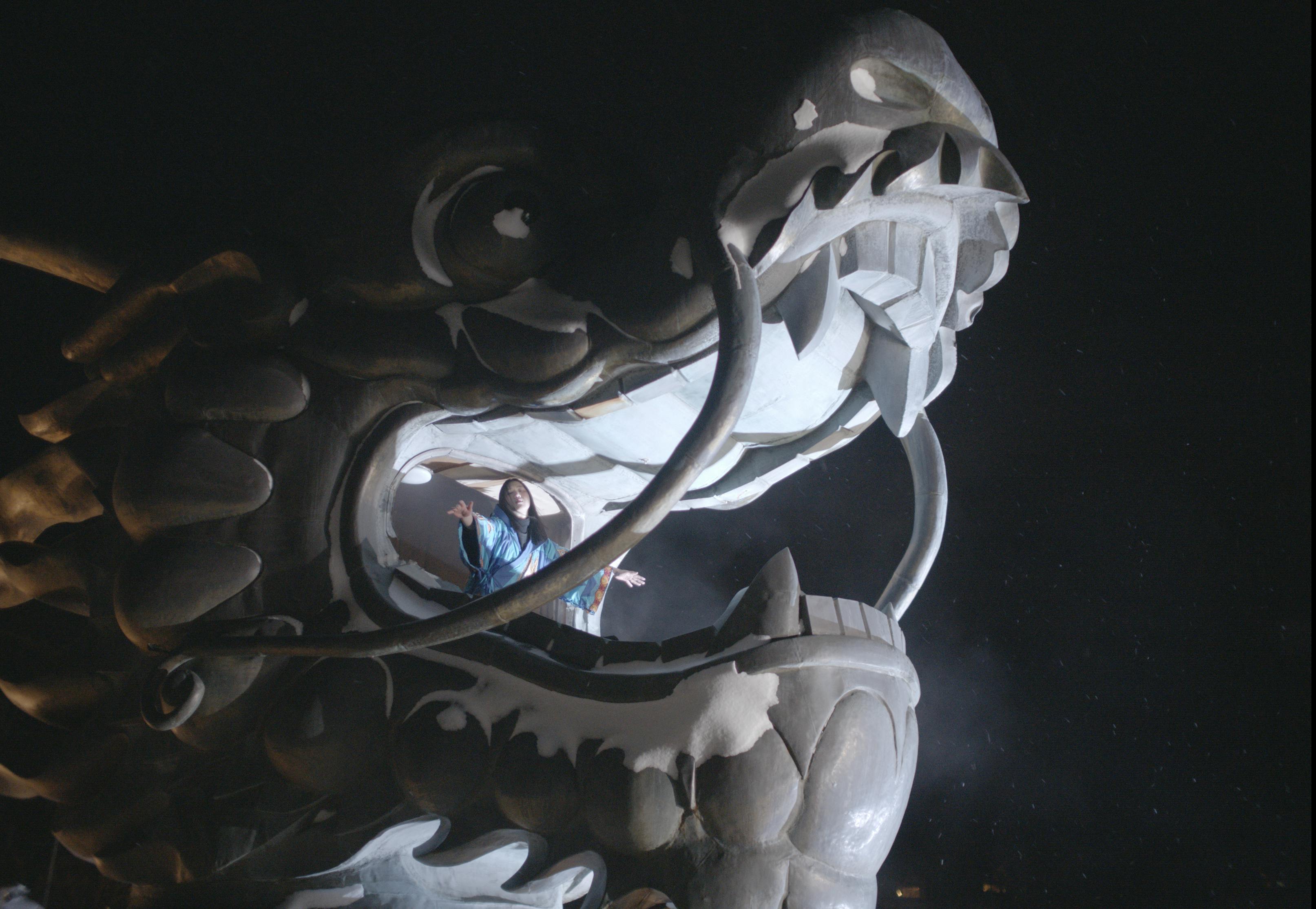 An Asian woman blue embroidered kimono stands with outstretched arms within the open mouth of a Chinese dragon sculpture. The image is taken from a film still. The still has been captured at night, and the dragon is lit from below.