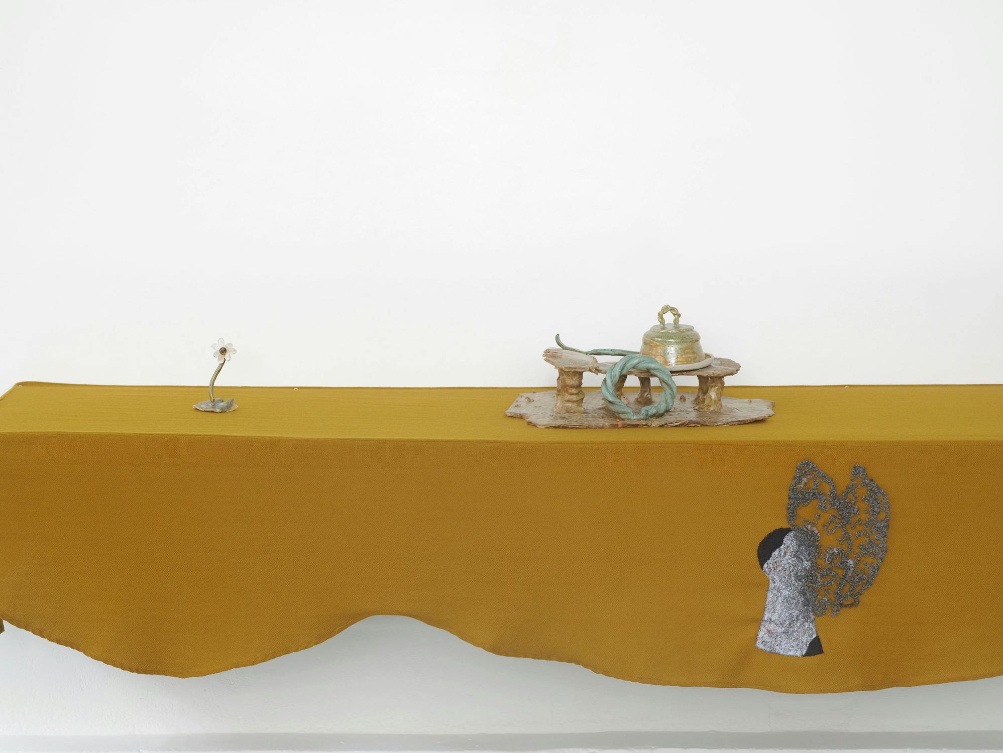 a wall-mounted shelf draped with mustard-coloured fabric. The shelf display a small ceramic flower and a larger glazed sculptural work.