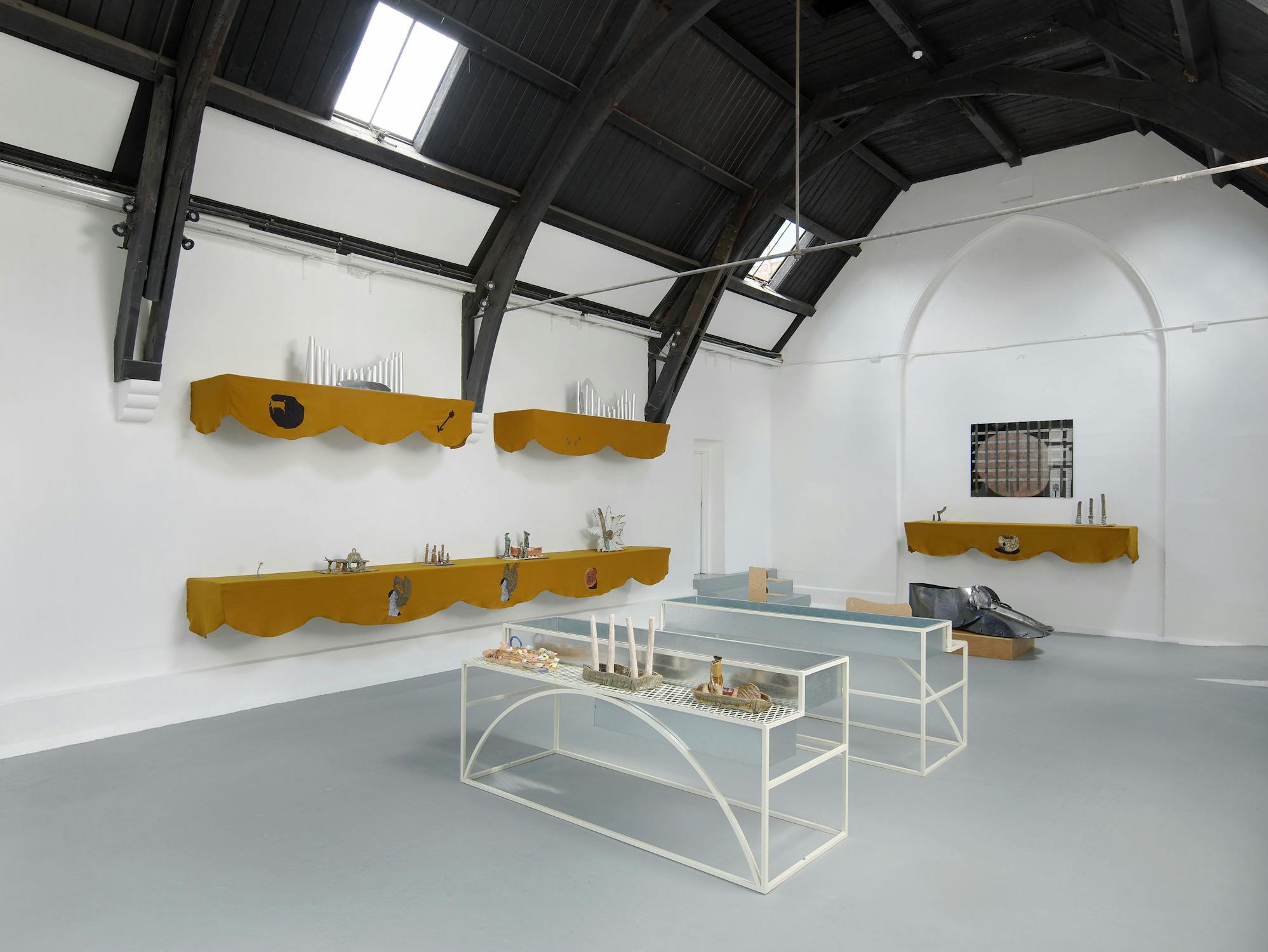 A gallery space with white walls and a grey floor. Shelves draped with mustard-coloured fabric are mounted on the walls, displaying various objects. Two white framed vitrines display different ceramic objects in the middle of the room.