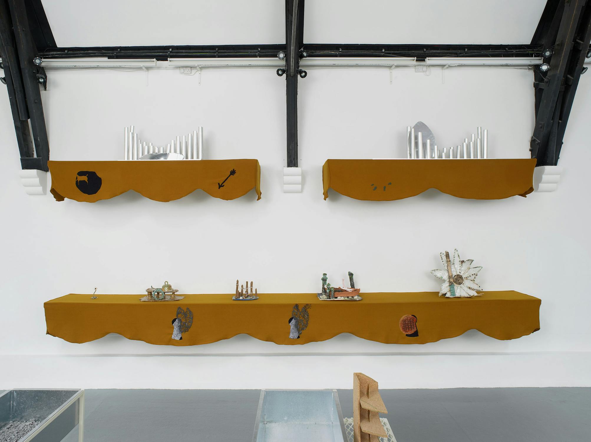 A view of an artwork installation comprising three wall-mounted shelves draped with mustard-coloured fabric. The shelves display various small artworks and objects.