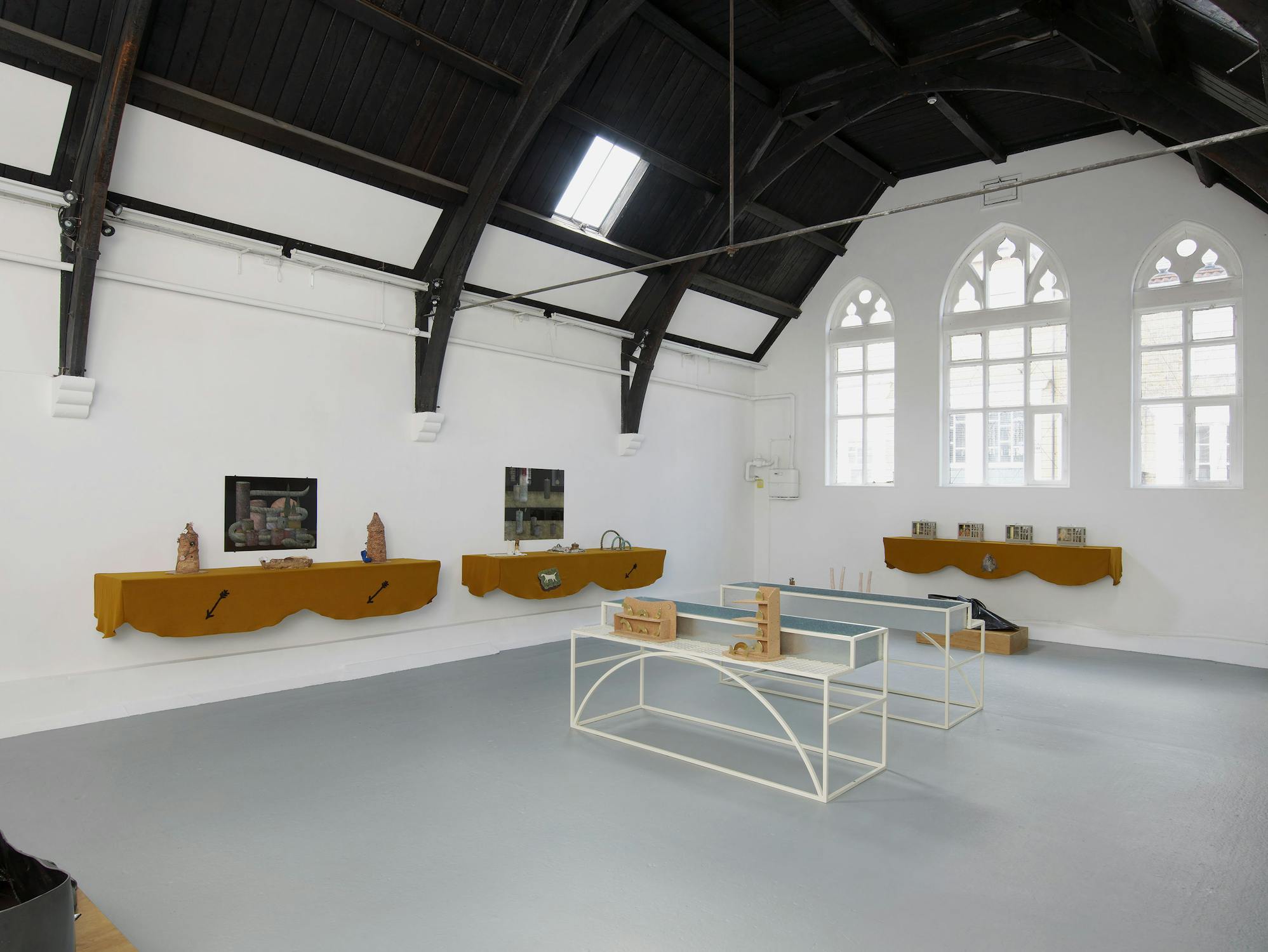 A gallery space with white walls, three large chapel windows and a grey floor. Shelves draped with brown fabric are mounted on the walls, displaying various objects. In the middle of the room, two white framed vitrines display different ceramic objects.