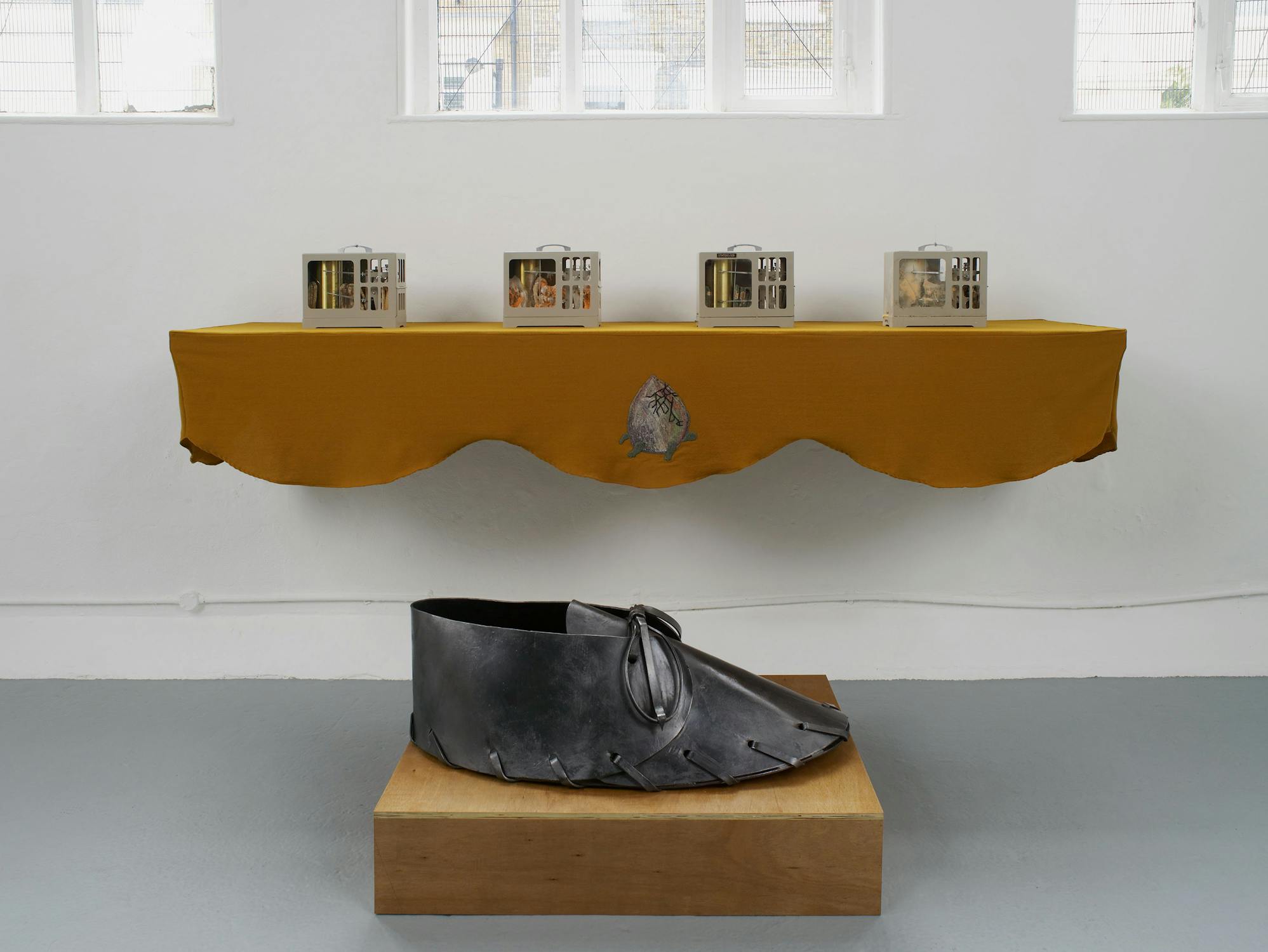 A view of an artwork installation comprising a wall-mounted shelf draped with mustard-coloured fabric. The display comprises four ceramic artworks. An artwork of an oversized leather shoe sits in the foreground