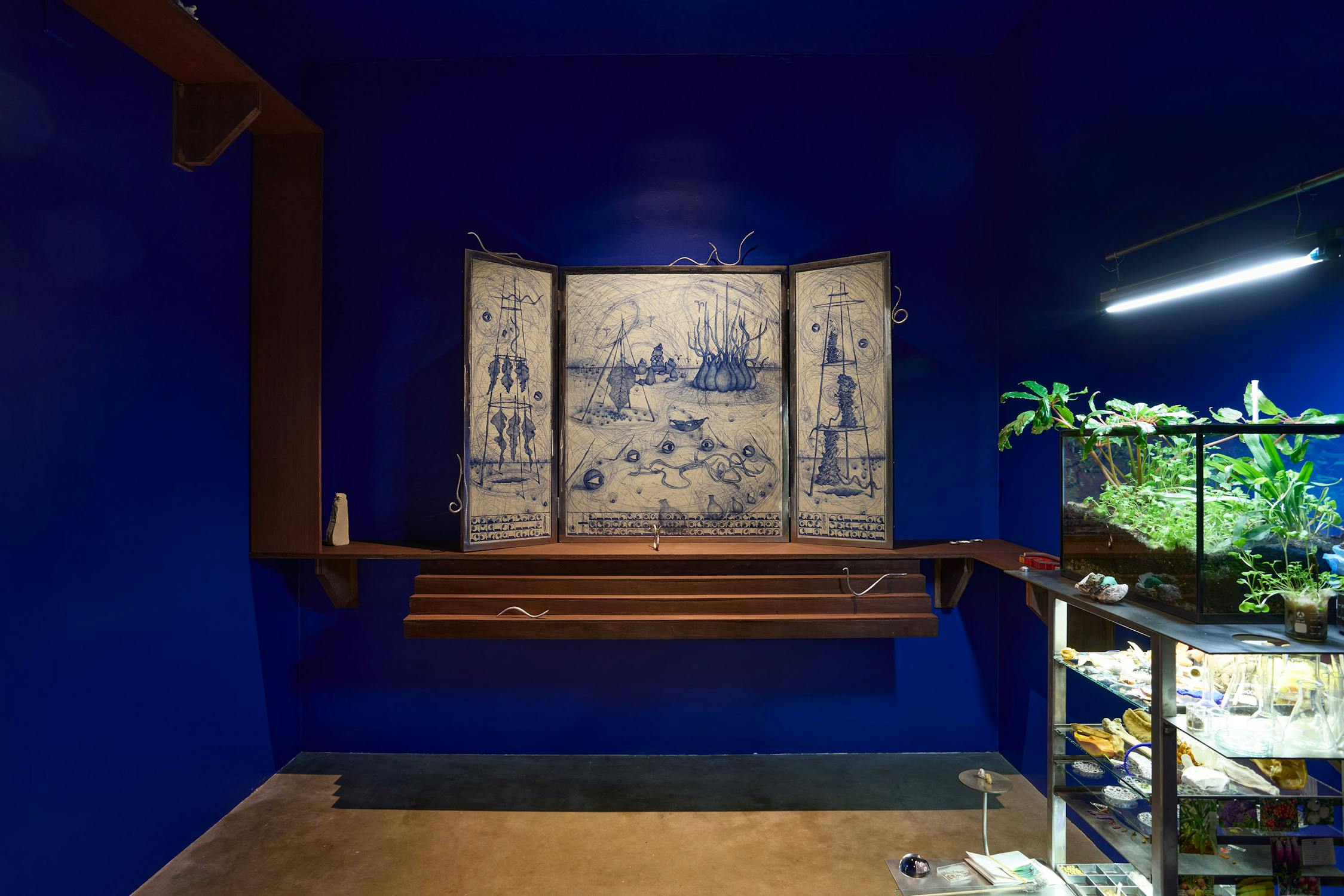 An installation including a triptych frame containing a drawing in blue ink installed on a wooden shelf with miniature steps