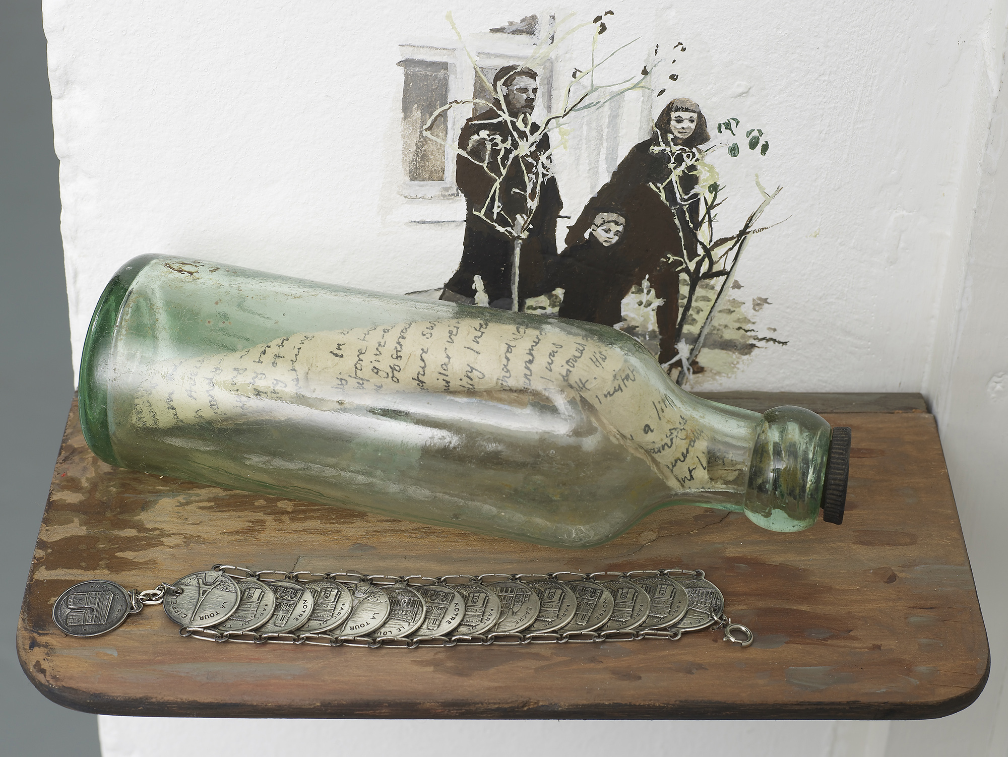 A glass bottle with a handwritten letter inside rests on a wooden shelf, next to a coin bracelet. Behind them is a wall painting of three people, including a child, holding thin branches.