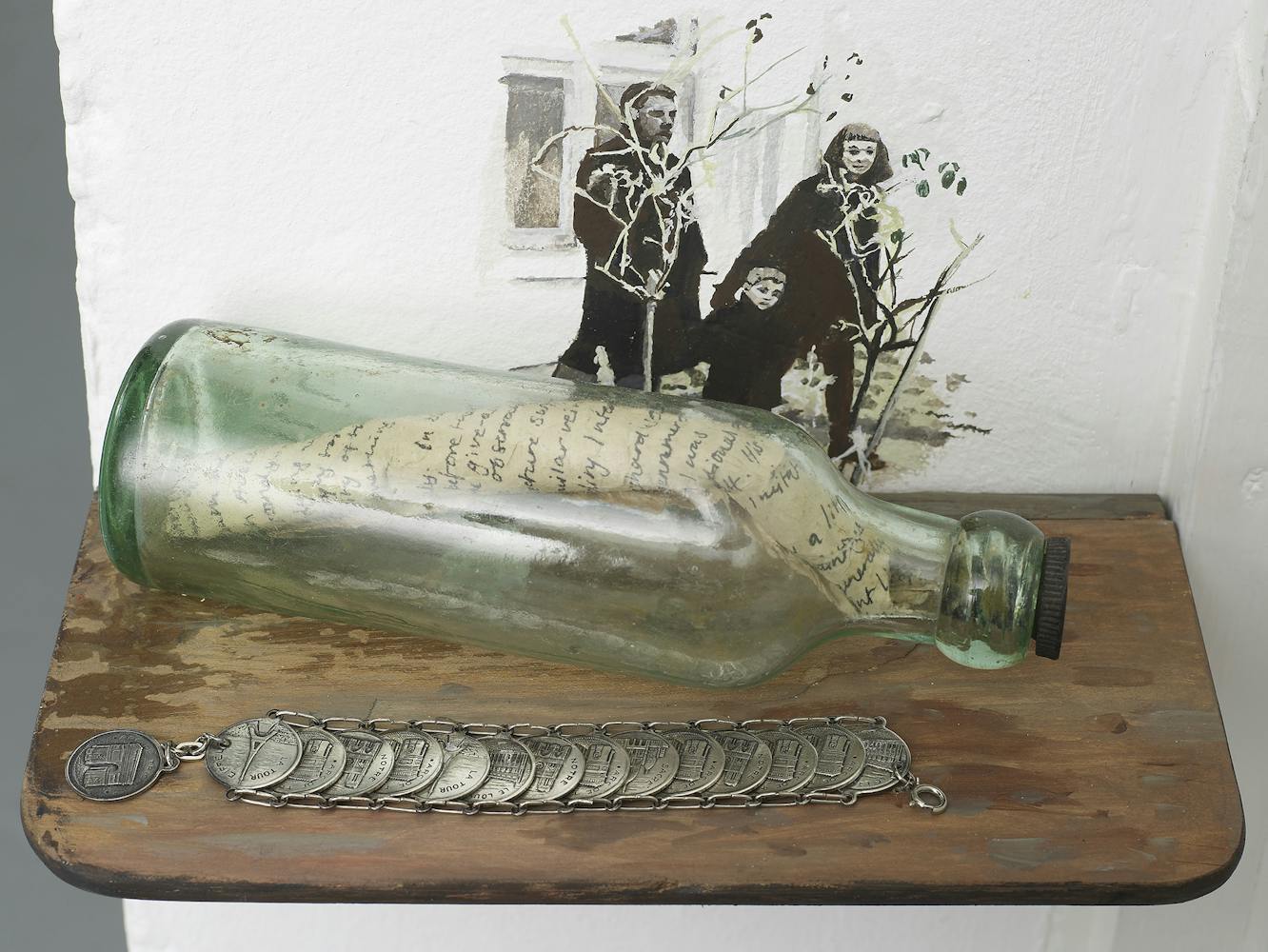 A glass bottle with a handwritten letter inside rests on a wooden shelf, next to a coin bracelet. Behind them is a wall painting of three people, including a child, holding thin branches.