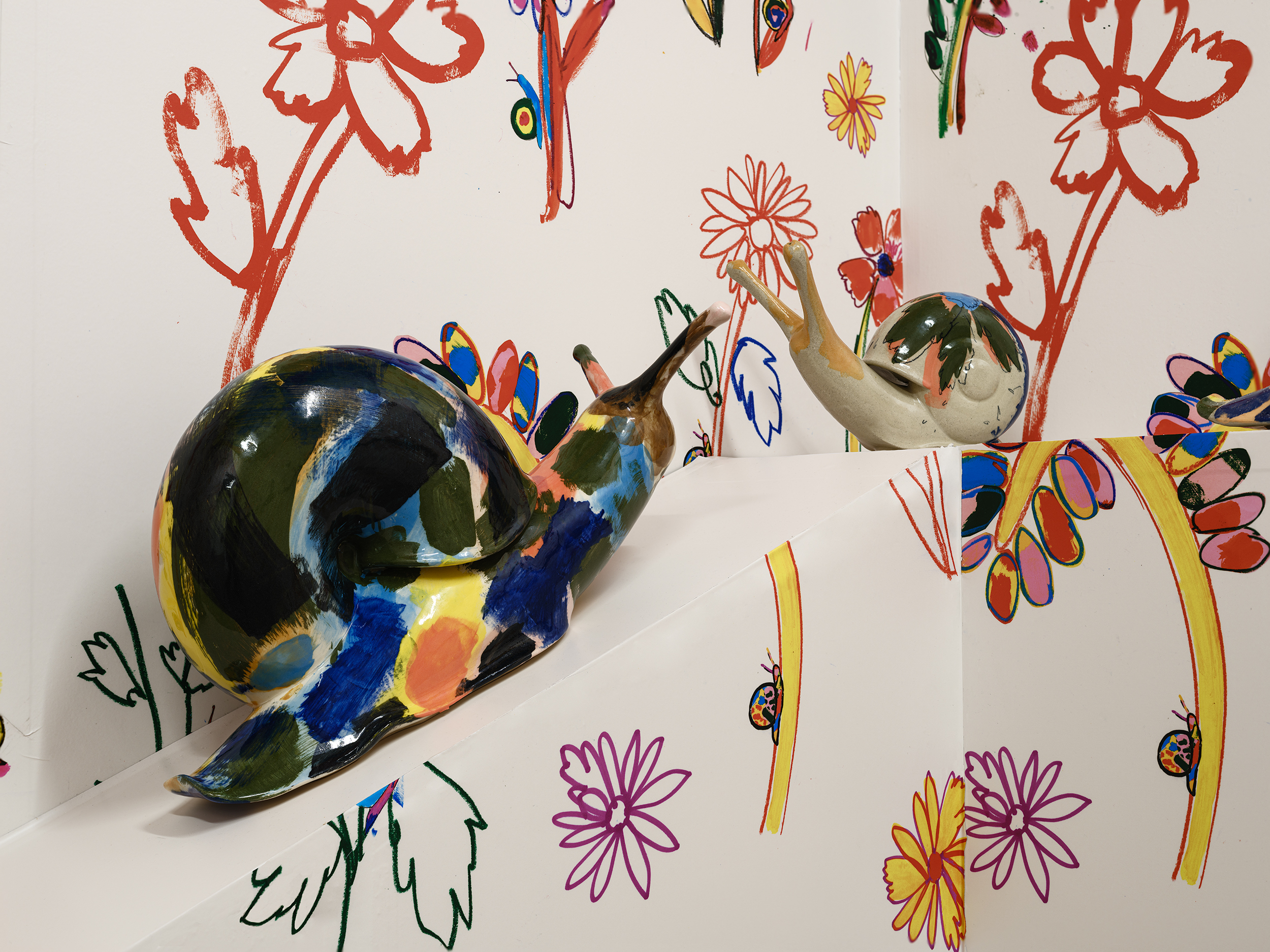 Two colourful glazed ceramic snail sculptures face each other on a white ledge, with vibrant, hand-drawn flowers in red, yellow, and purple covering the white walls behind them.