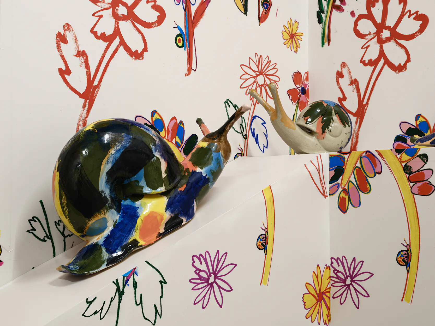 Two colourful glazed ceramic snail sculptures face each other on a white ledge, with vibrant, hand-drawn flowers in red, yellow, and purple covering the white walls behind them.