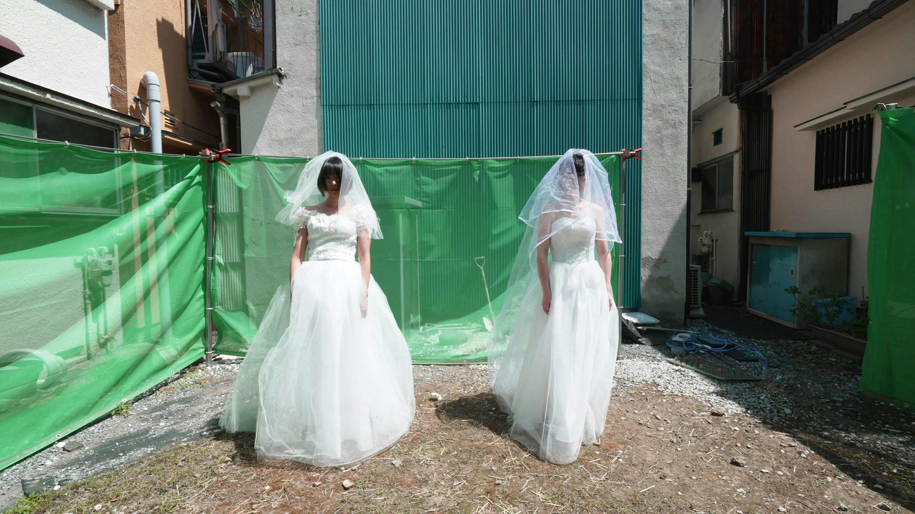 a film still that captures two women in identical bridal gowns with veils, standing in an outdoor space, enclosed by industrial buildings