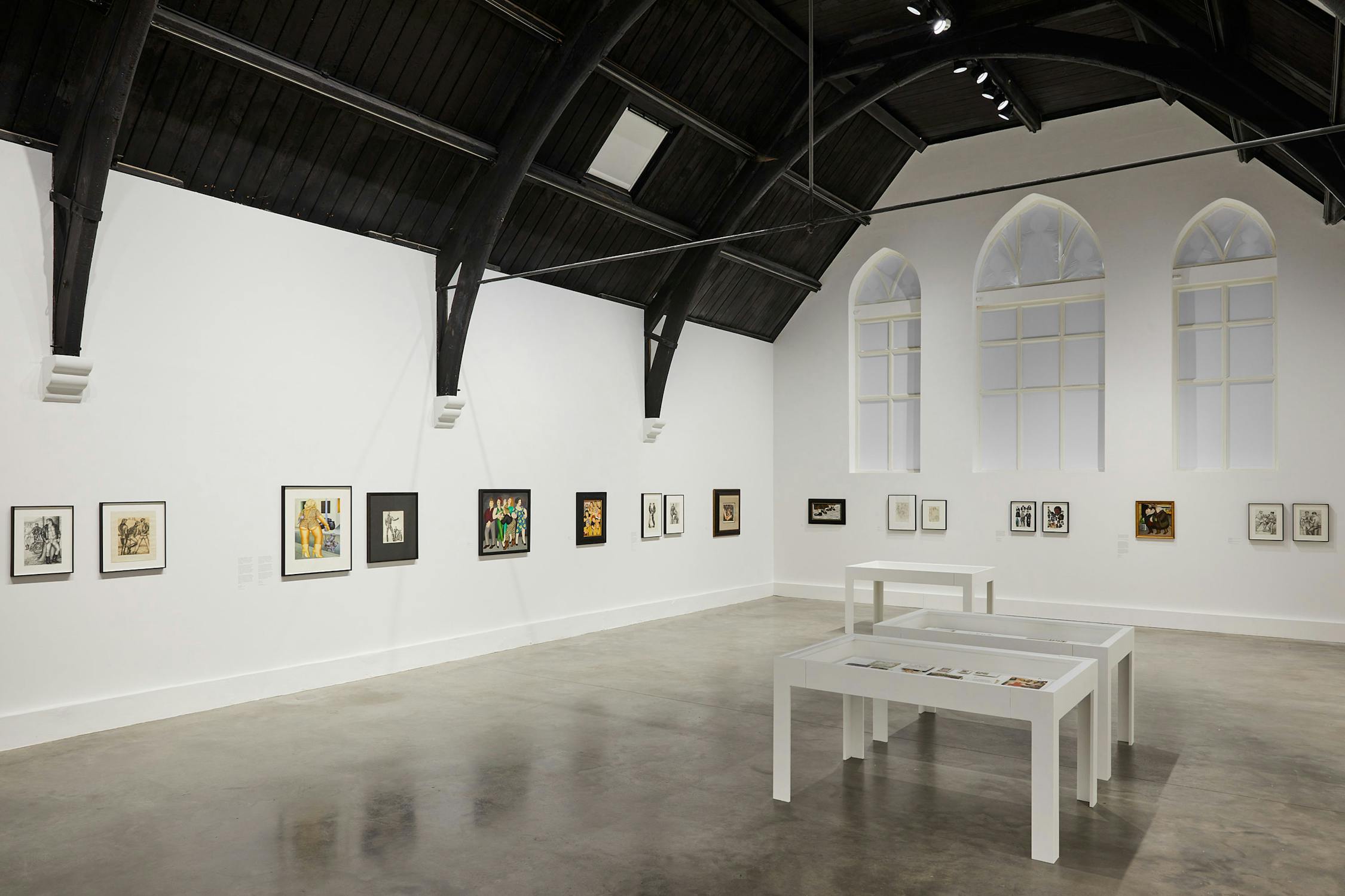 a gallery with white walls, dark vaulted ceiling and polished concrete floors. Artworks by Beryl Cook and Tom of Finland line the walls, and three vitrines are positioned within the space.