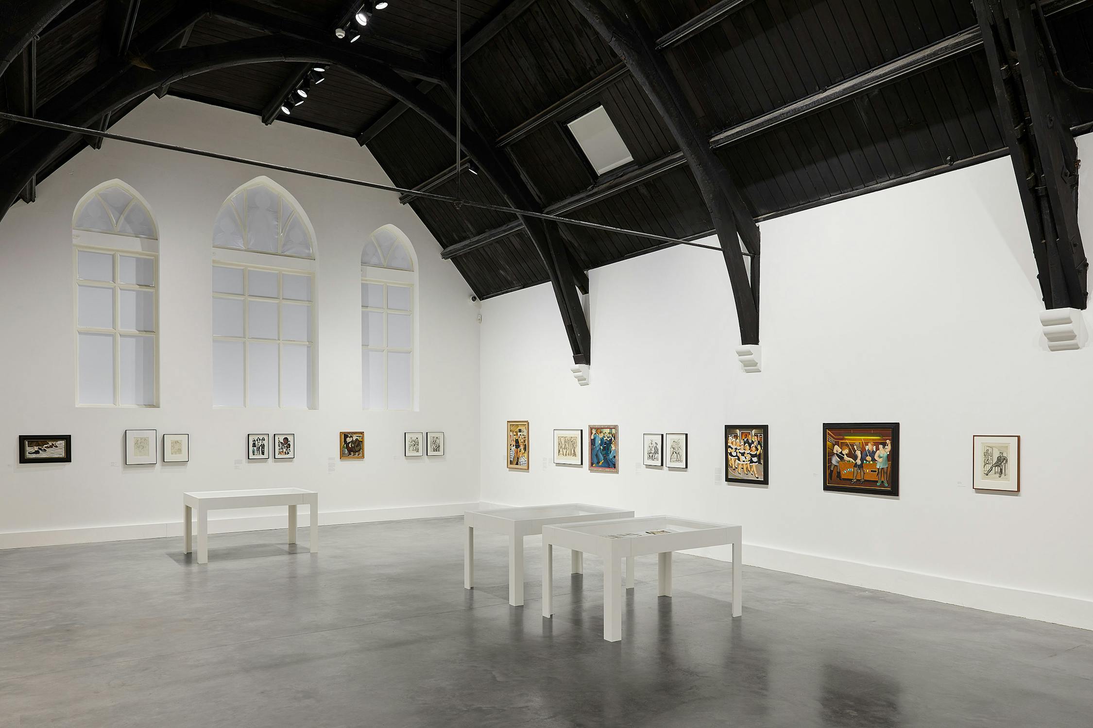 a brightly-lit gallery with white walls, dark vaulted ceiling and polished concrete floors. Artworks by Beryl Cook and Tom of Finland line the walls, and three vitrines are positioned within the space.