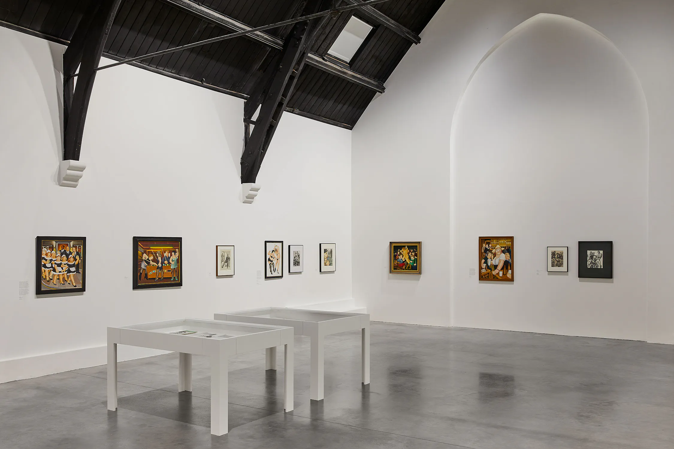 a brightly-lit gallery with white walls and polished concrete floors. Colourful-painted artworks by Beryl Cook and detailed-graphic pencil drawings by Tom of Finland line the walls, and three vitrines are positioned within the space.