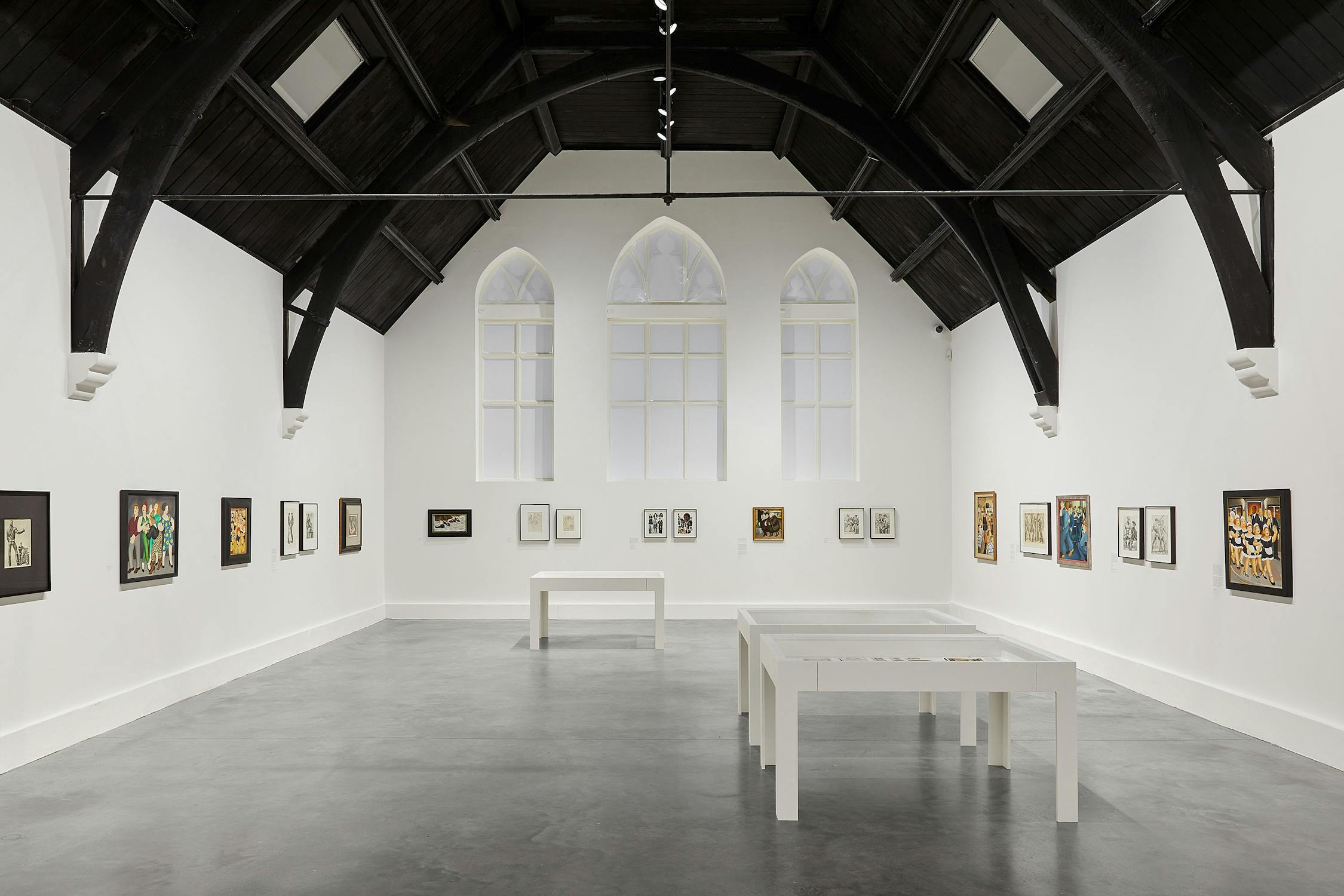 a brightly-lit gallery with white walls, dark vaulted ceiling and polished concrete floors. Artworks by Beryl Cook and Tom of Finland line the walls, and three vitrines are positioned within the space.