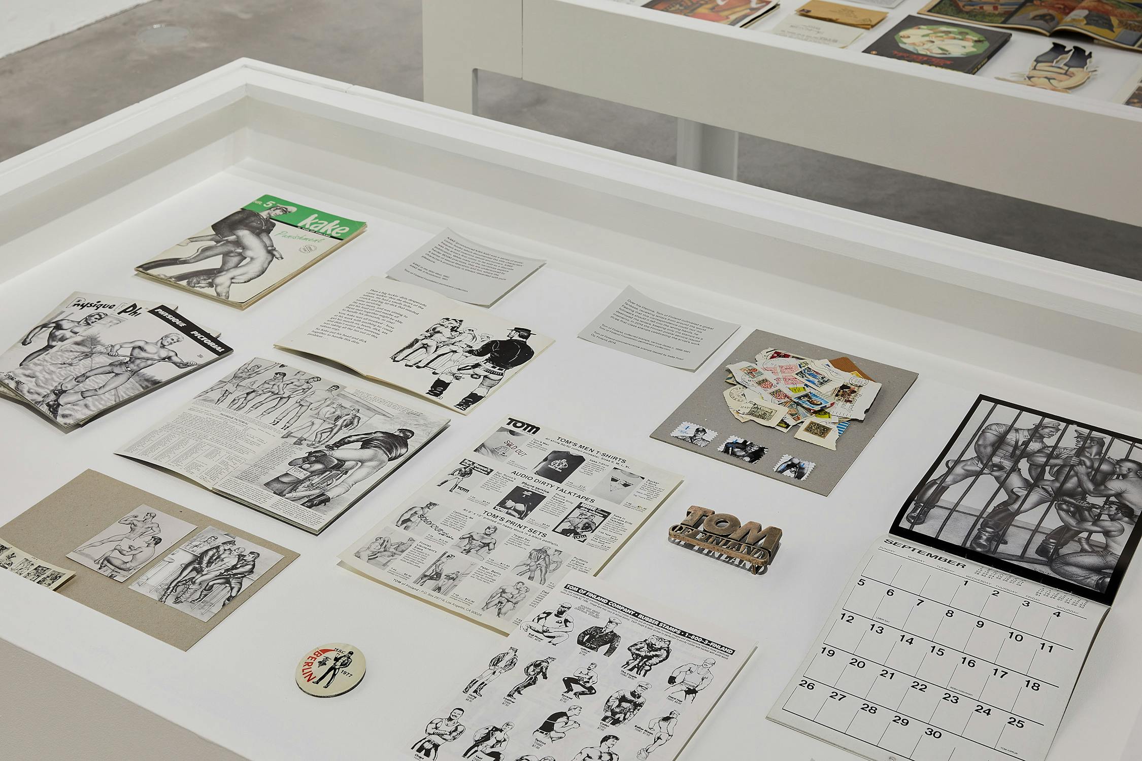 a white vitrine with glass top displaying memorabilia including calendars, badges, rubber0-stamp designs and postcards belonging to artist Beryl Cook