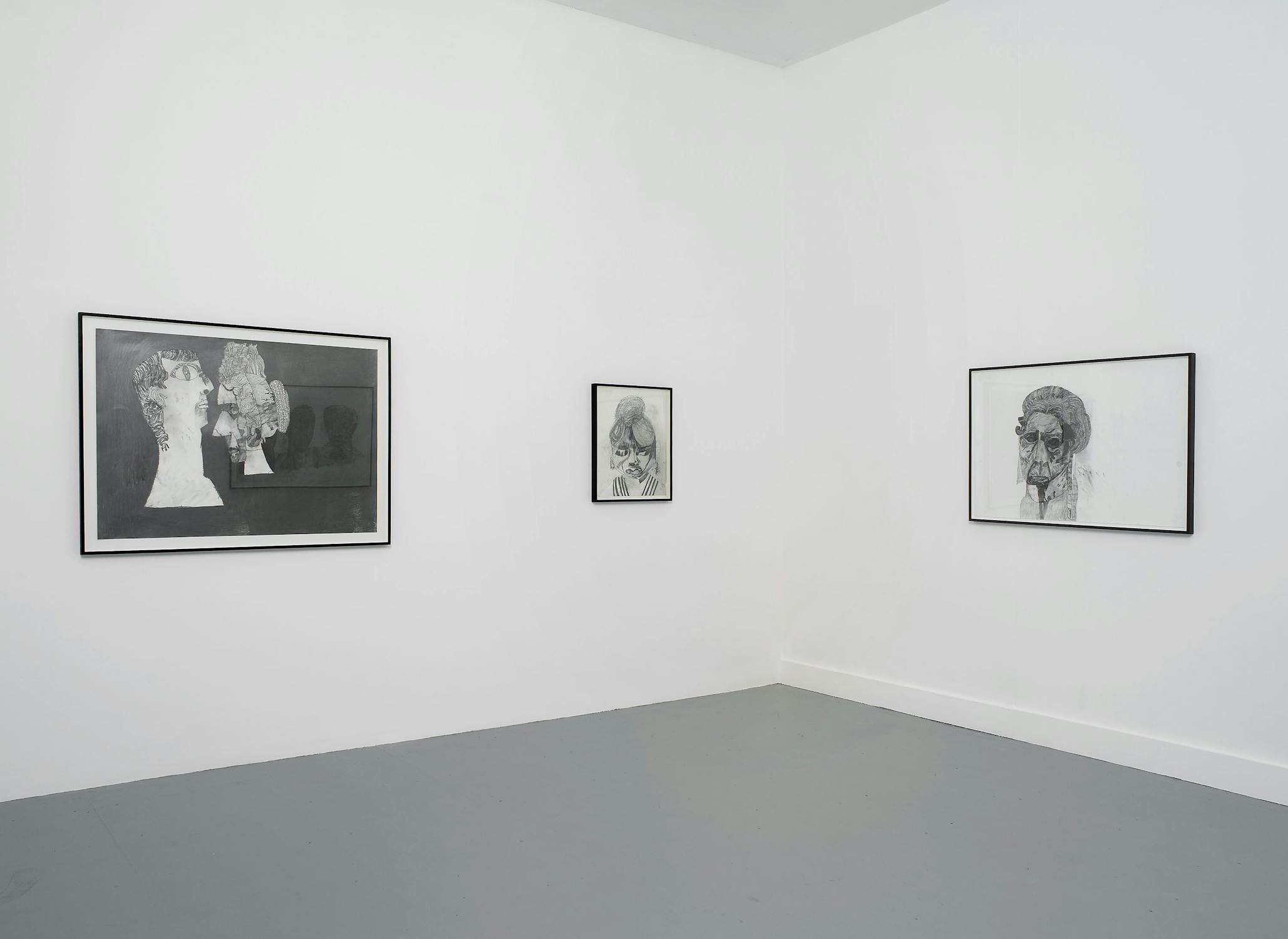 Three pencil drawings of figures with exaggerated features on white paper with black frames, hung in the corner of a white-walled gallery