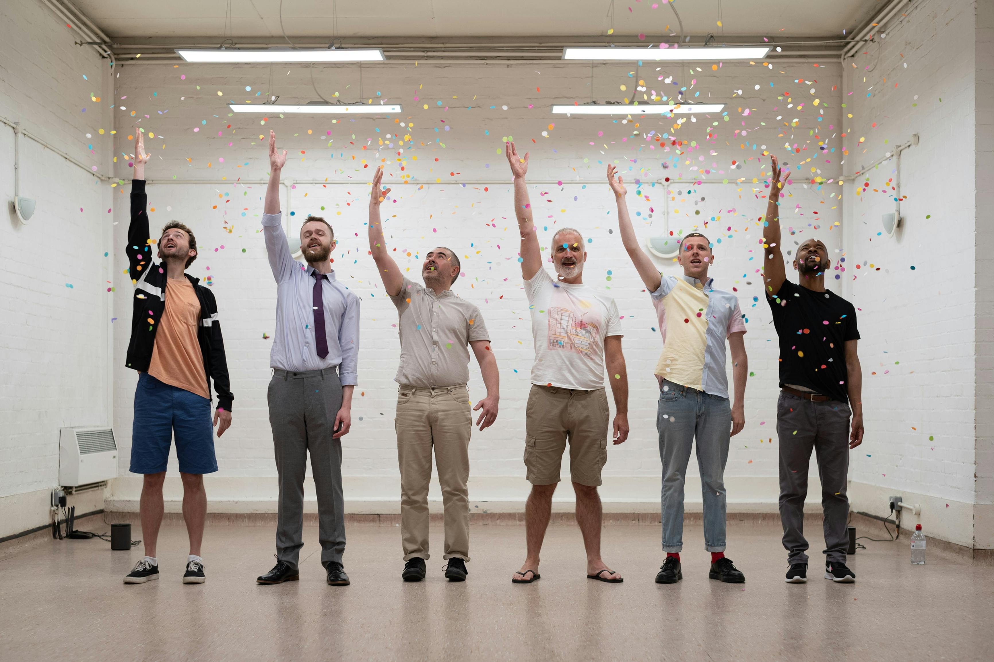 six men of varying ages and ethnicities stand in a line, each throwing confetti up in the air woth their right hands