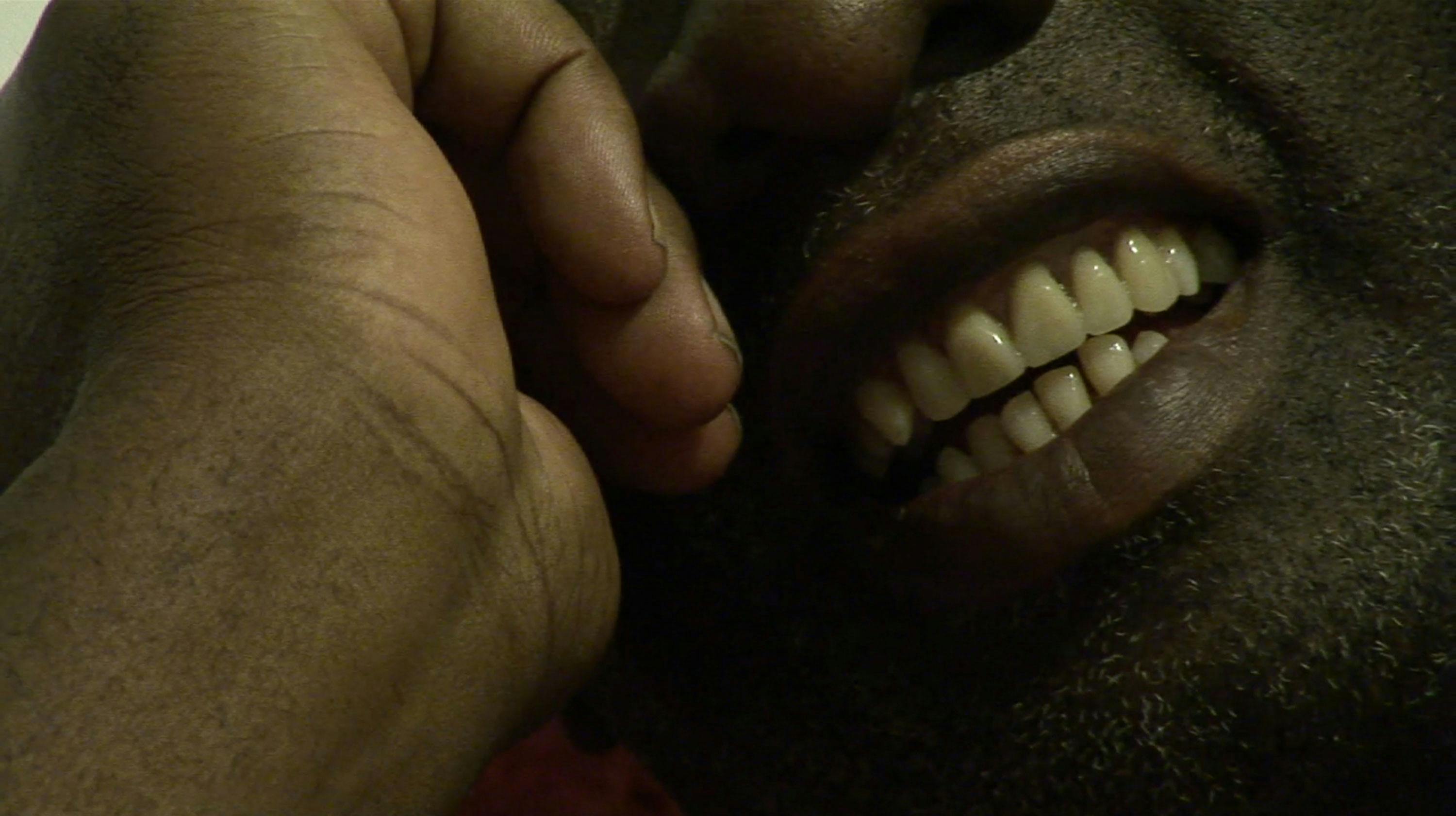 Close-up of a person smiling with their hand resting against their face. The image focuses on their mouth, showing clean, white teeth, and part of their nose and chin which is covered in stubble. The lighting appears dim.