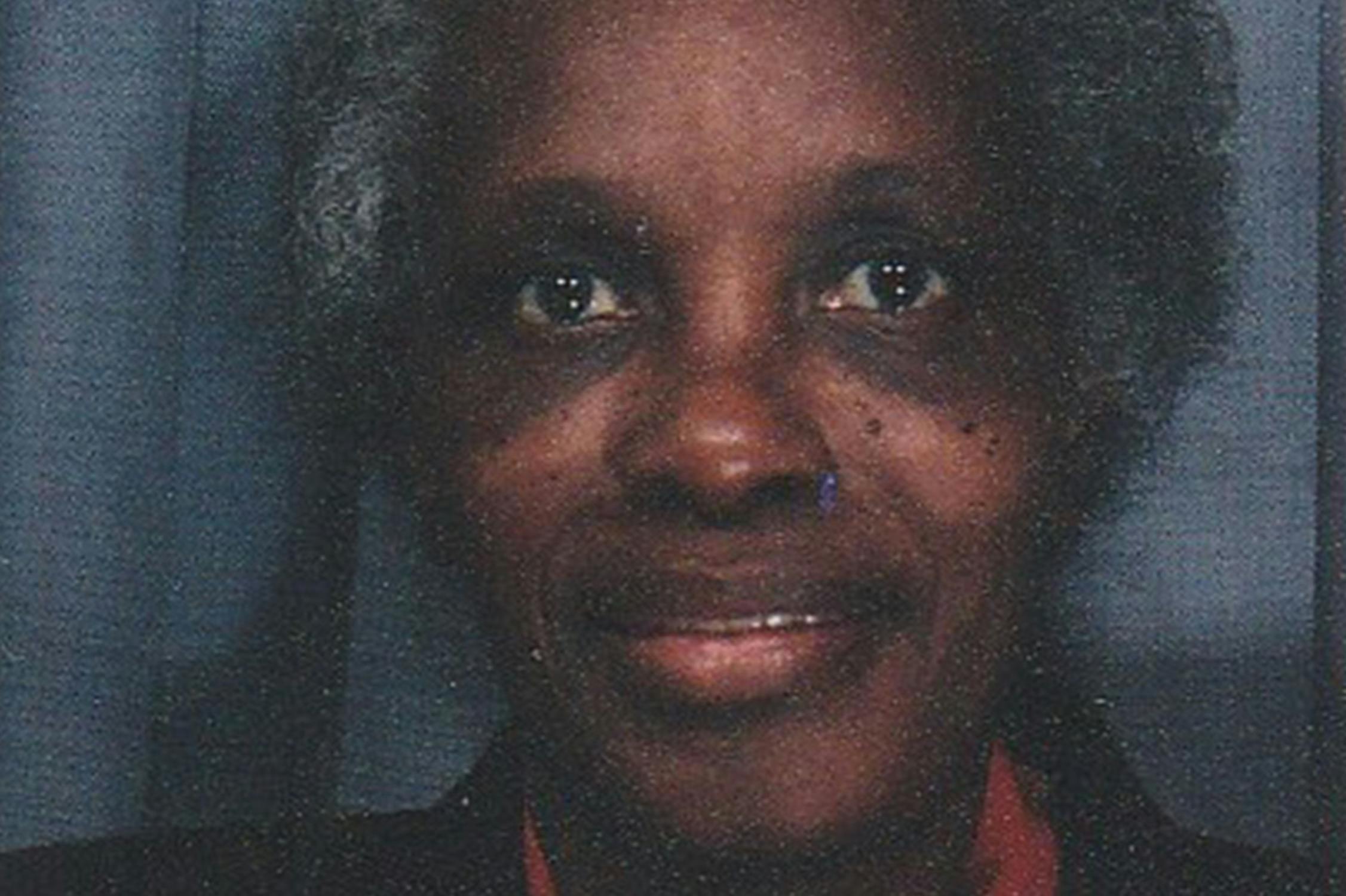 A close-up photo of Ain Bailey's mother, an older person with short, curly grey hair. They are smiling subtly and have a warm expression. The background is a plain dark grey curtain. The person is wearing a dark-coloured top.