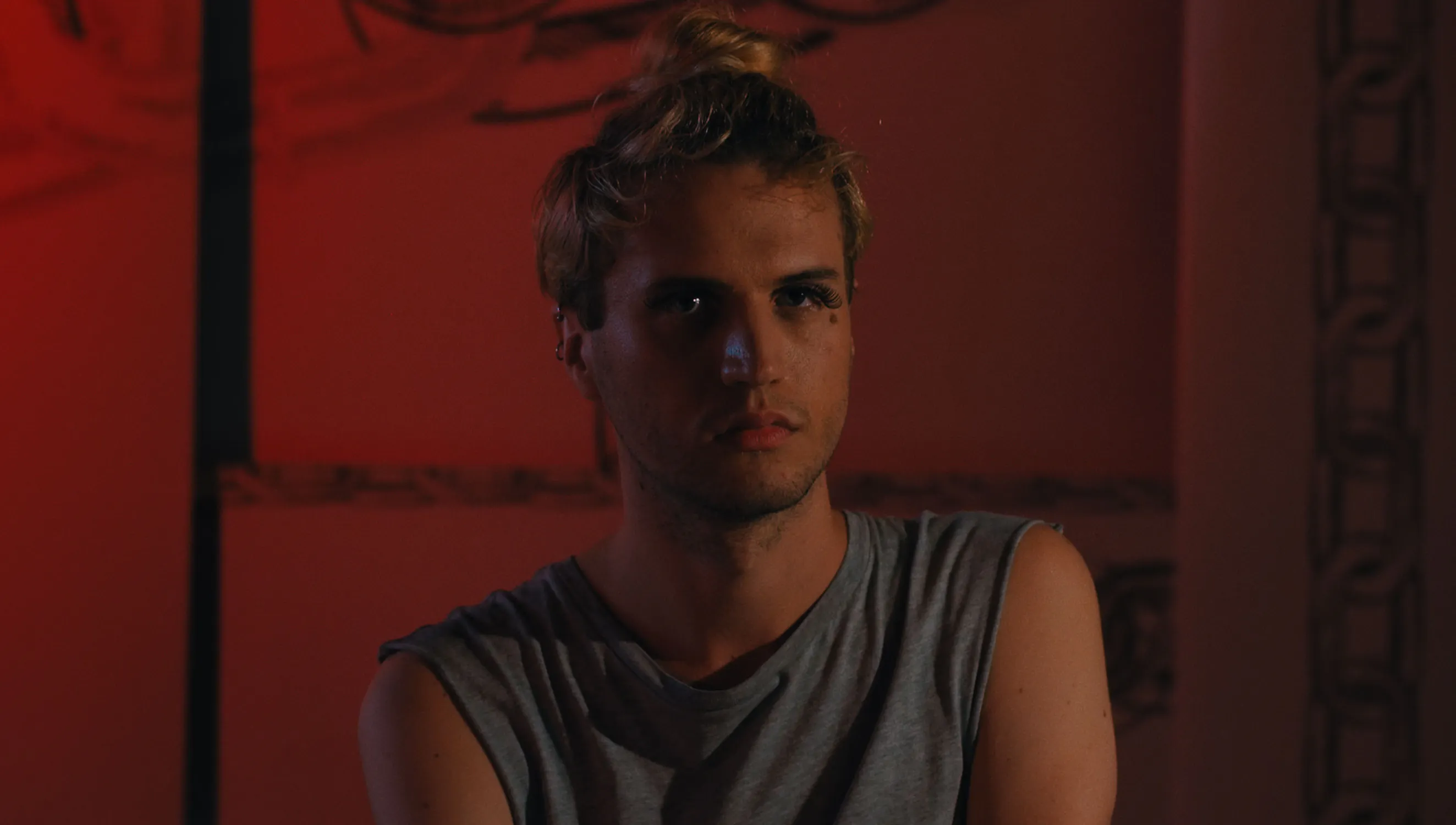 A person with shoulder-length blond hair tied in a bun is wearing a sleeveless gray shirt. The individual has a serious expression and is set against a dimly lit background with red lighting and some ornate wall details.
