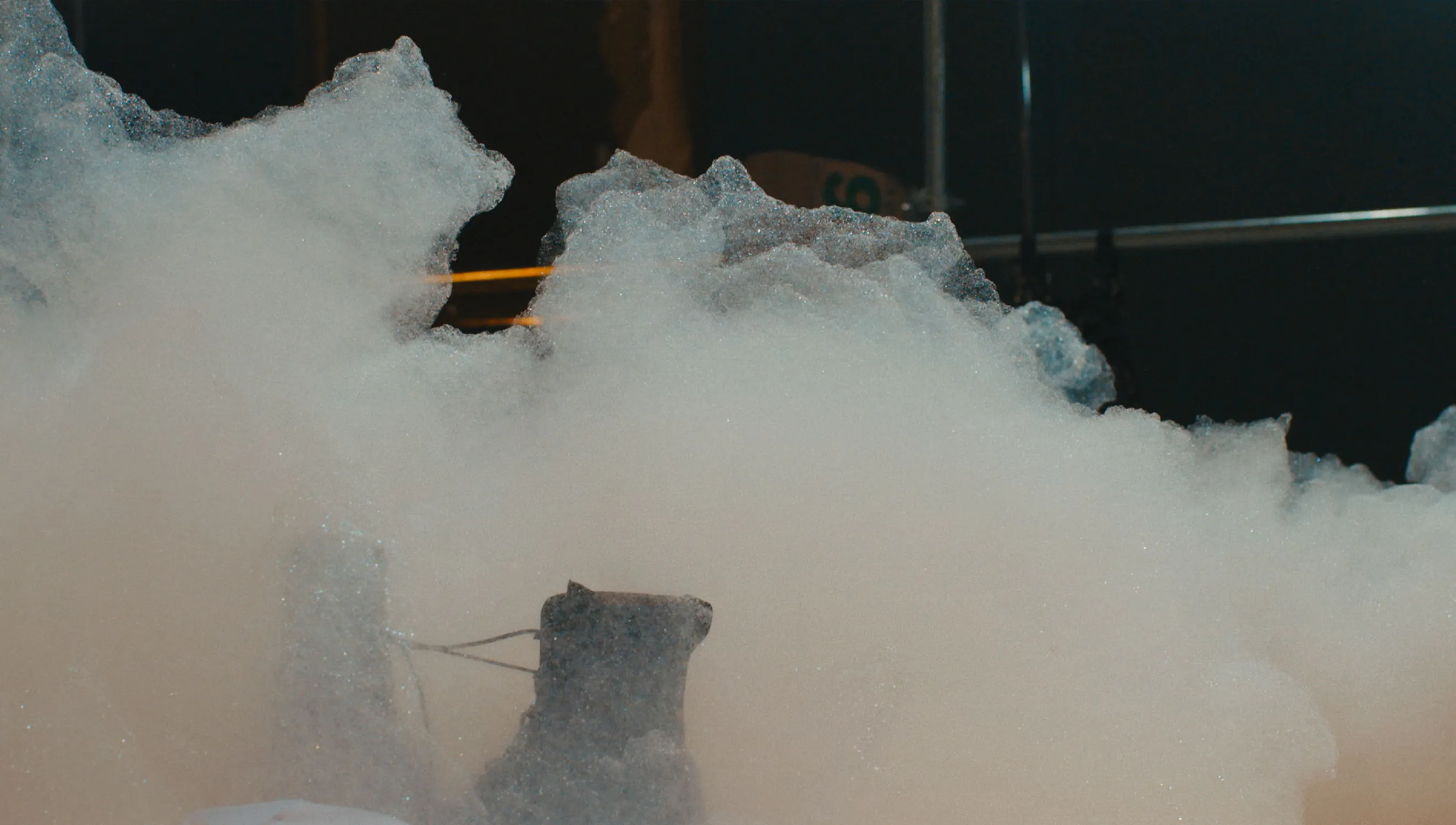 A dense cloud of white foam fills the scene, partially obscuring some objects and structures behind it. The foam appears to be thick and bubbly, with a hazy, indistinct background. There are glimpses of metallic elements and possibly a cardboard box.