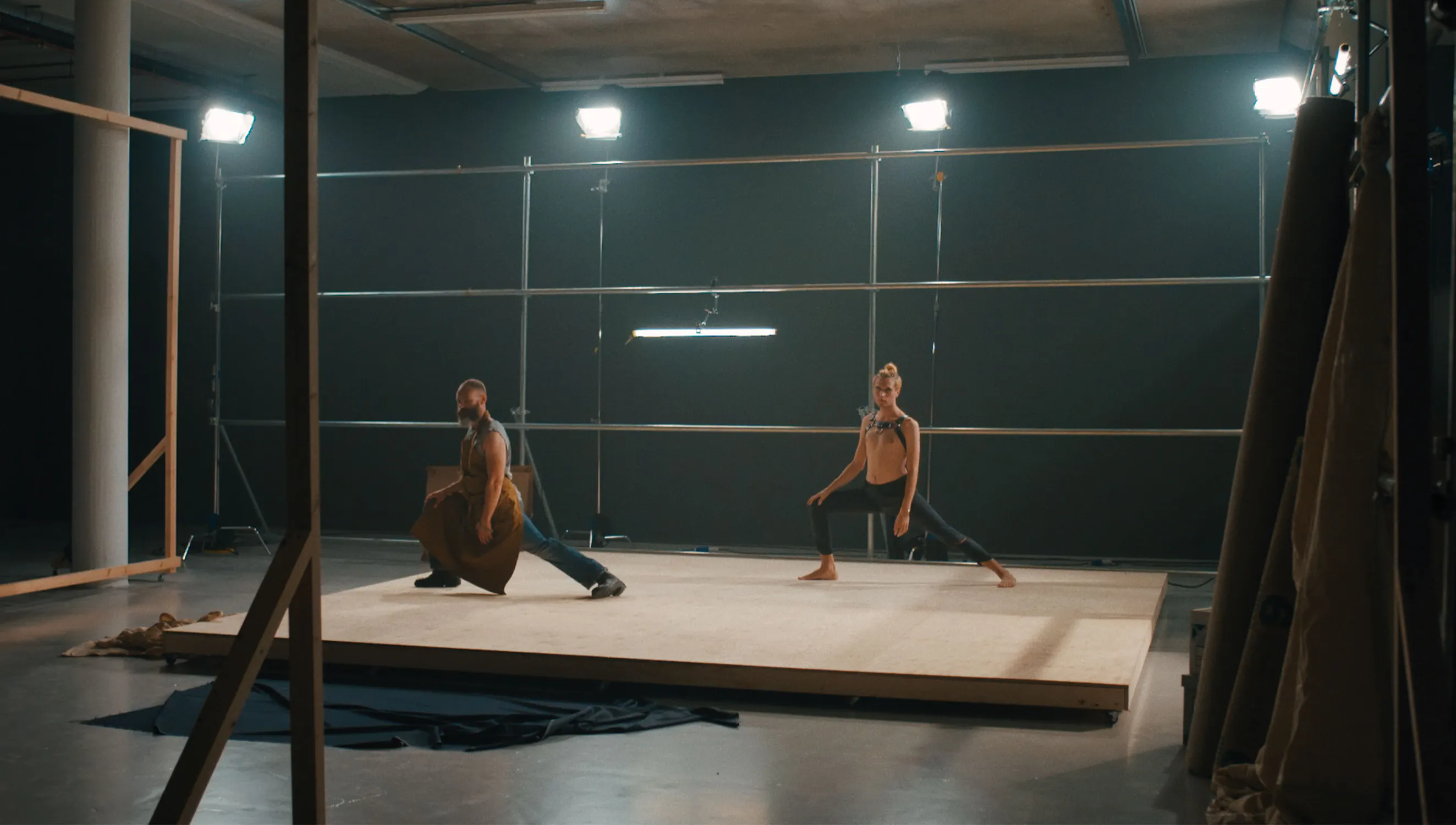 Two people are performing dance moves on a raised wooden platform in a studio with scaffolding and overhead lights. One dancer holds a brown cloth while both strike a pose, stretching one leg out and leaning forward. The background and floor are industrial in appearance.