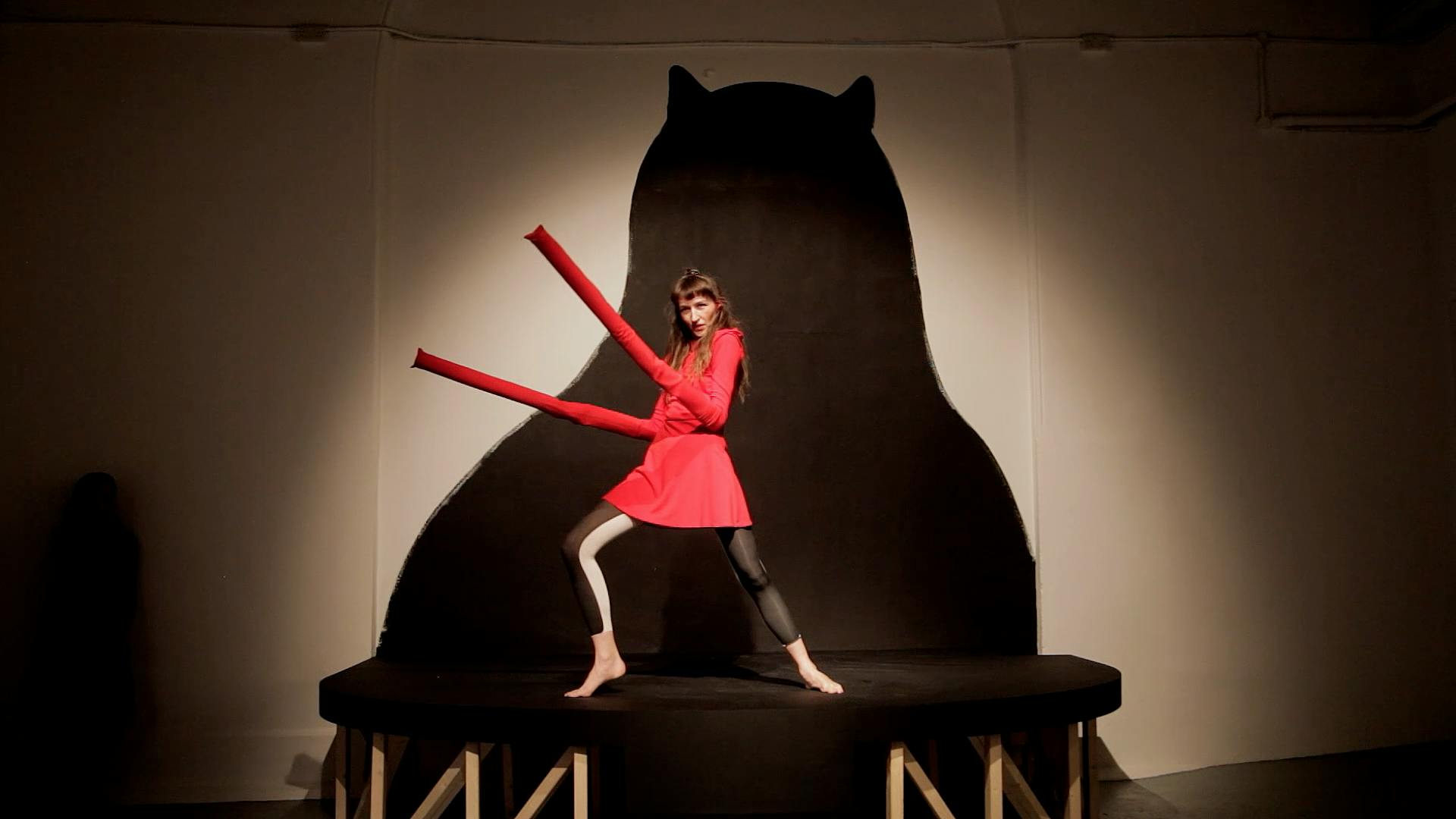 A person striking a dynamic pose on a small stage, wearing a bright red outfit and holding long red tubes. Behind them is a large black silhouette of a bear, on the wall, highlighted by focused lighting.