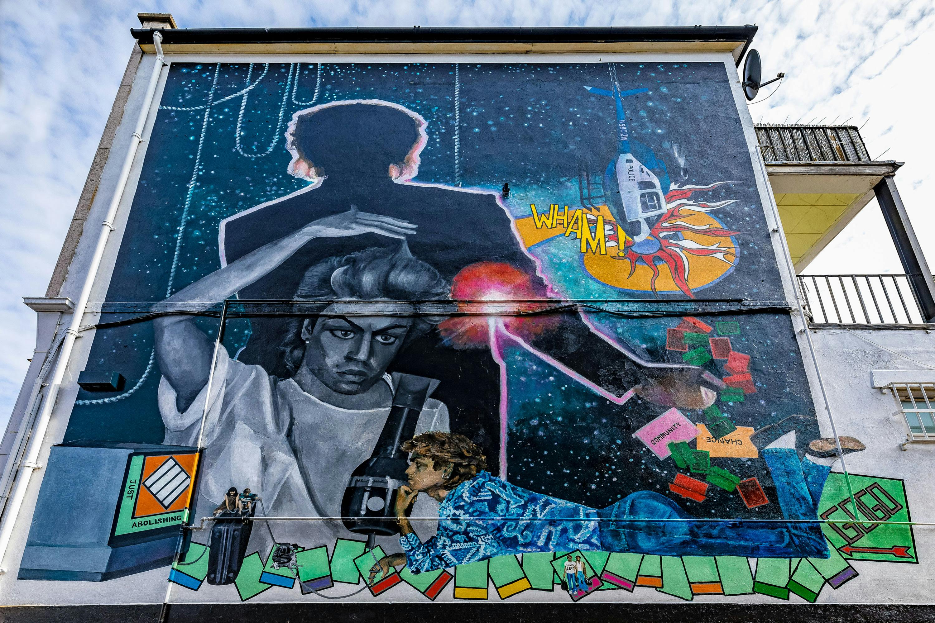 A large, colourful mural covers the side of a building. The artwork features singer George Michael with various objects around him, including a rocket, a Rubik's cube, and a vintage TV. The background is space-themed with stars and a cosmic vibe. Text in the image reads "WHAT AM I?.