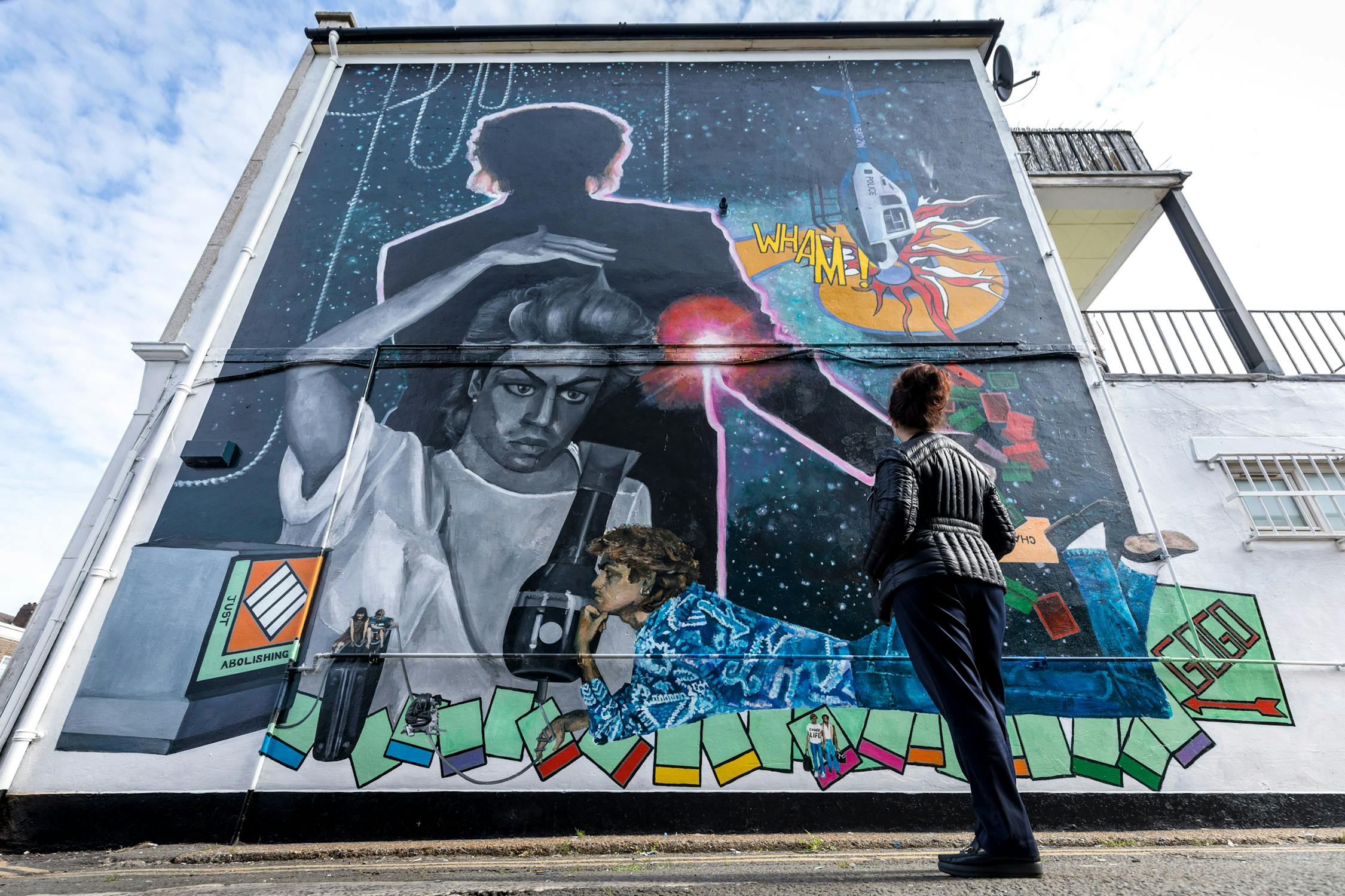 A person stands before a large, colourful mural painted on the side of a building. The mural depicts various abstract and cosmic elements, including singer George Michael, geometric shapes, and vibrant colours. A person gazes up at the artwork.