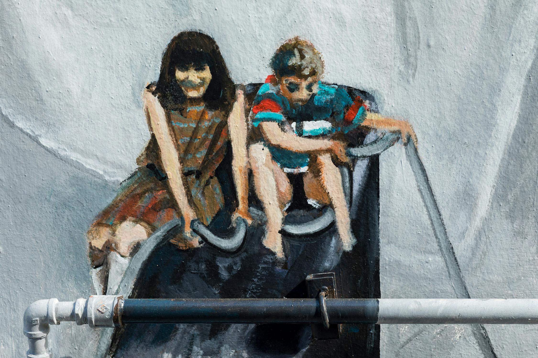 A mural depicts two figures sitting on a slide. The person on the left wears a striped dress, and the person on the right, a striped shirt and shorts. A metal pipe runs horizontally across the bottom of the mural in the foreground.