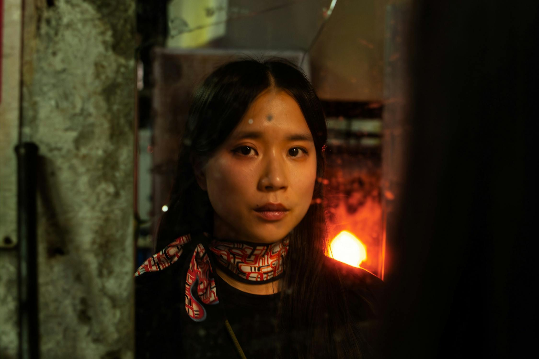 Portrait of artist Lap-See Lam wearing a patterned scarf. She is standing in front of a reflective surface, with a warm, glowing light behind her, creating a dramatic atmosphere.