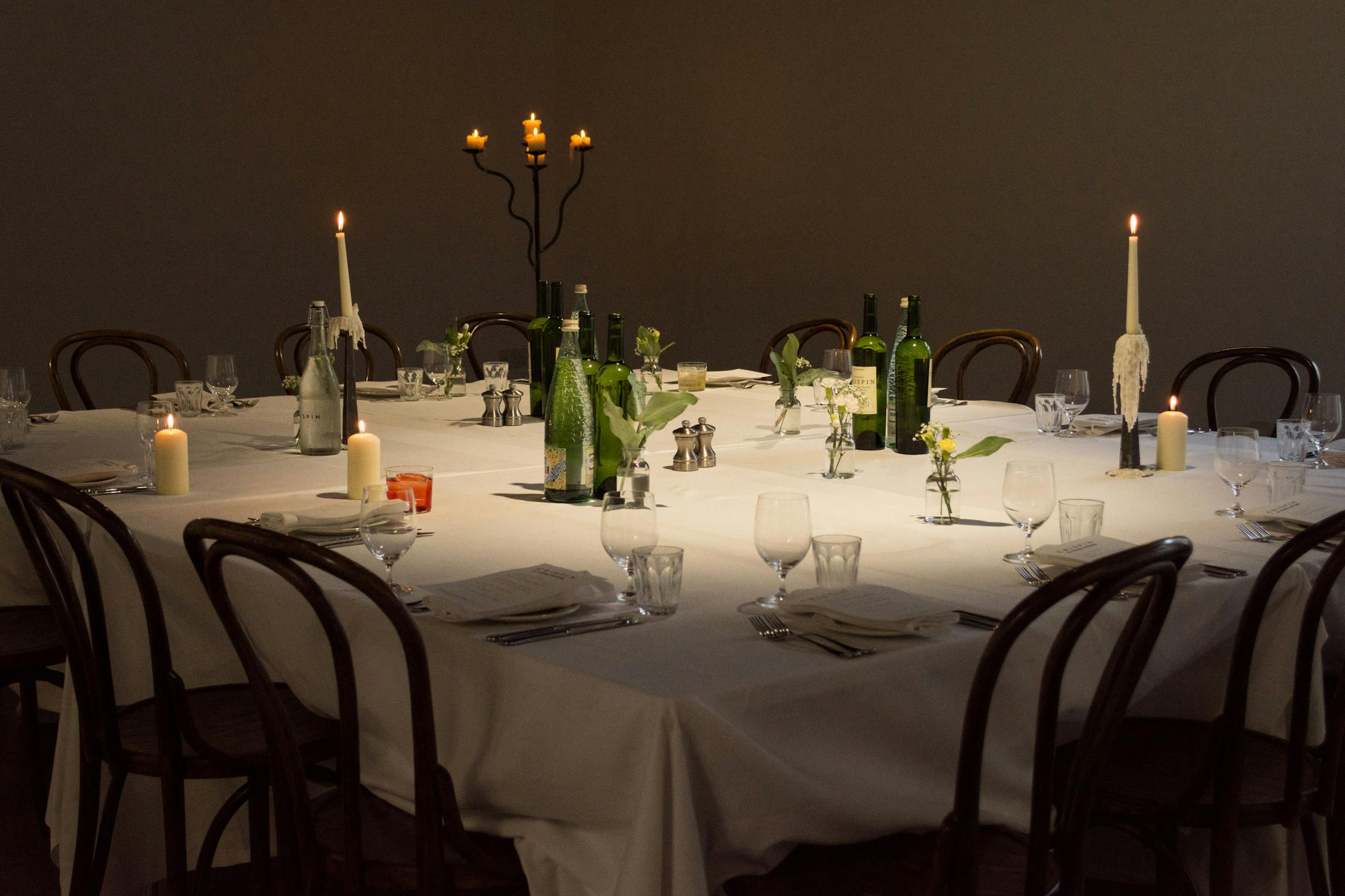 A large square dining table with a white tablecloth is elegantly set for a meal. It features wine bottles, glassware, white napkins, and floral centerpieces. Candles, including a candelabra, create a warm, inviting atmosphere. Wooden chairs surround the table.