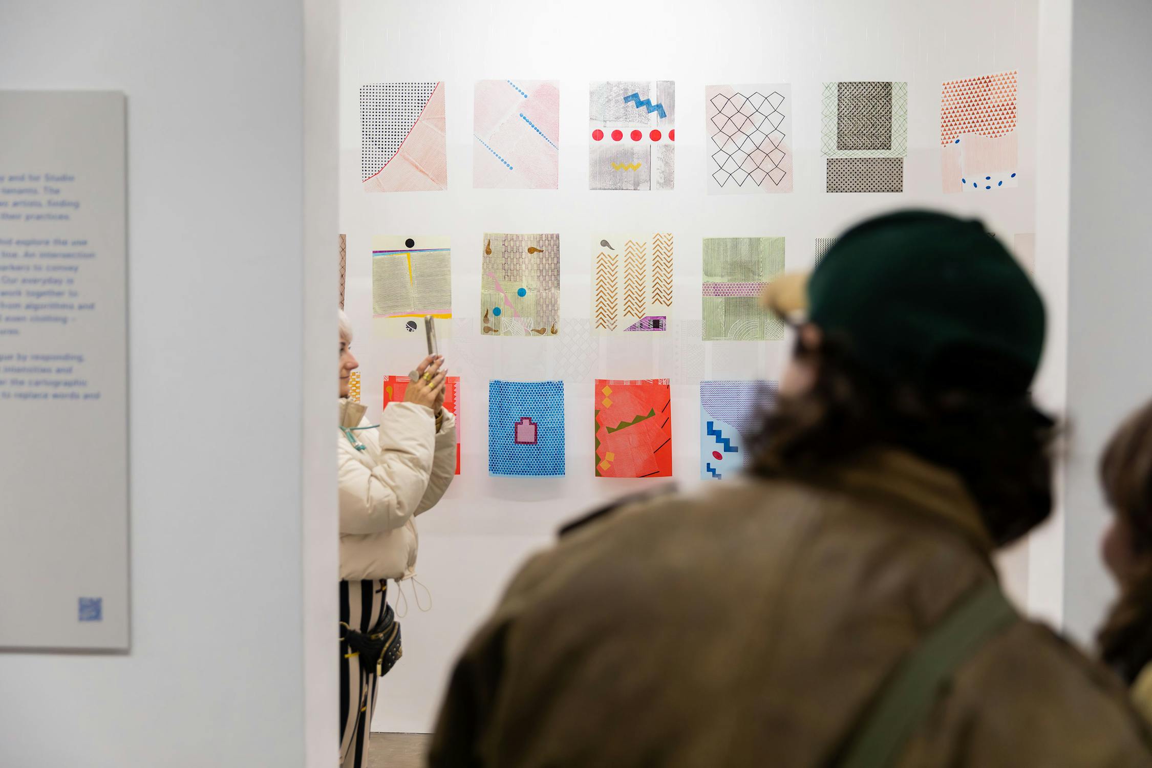 A person in a green cap looks at an art display with another individual in a white jacket at a gallery. The display features a series of small, colourful, geometric prints on a white wall. A descriptive panel is partially visible on the left.