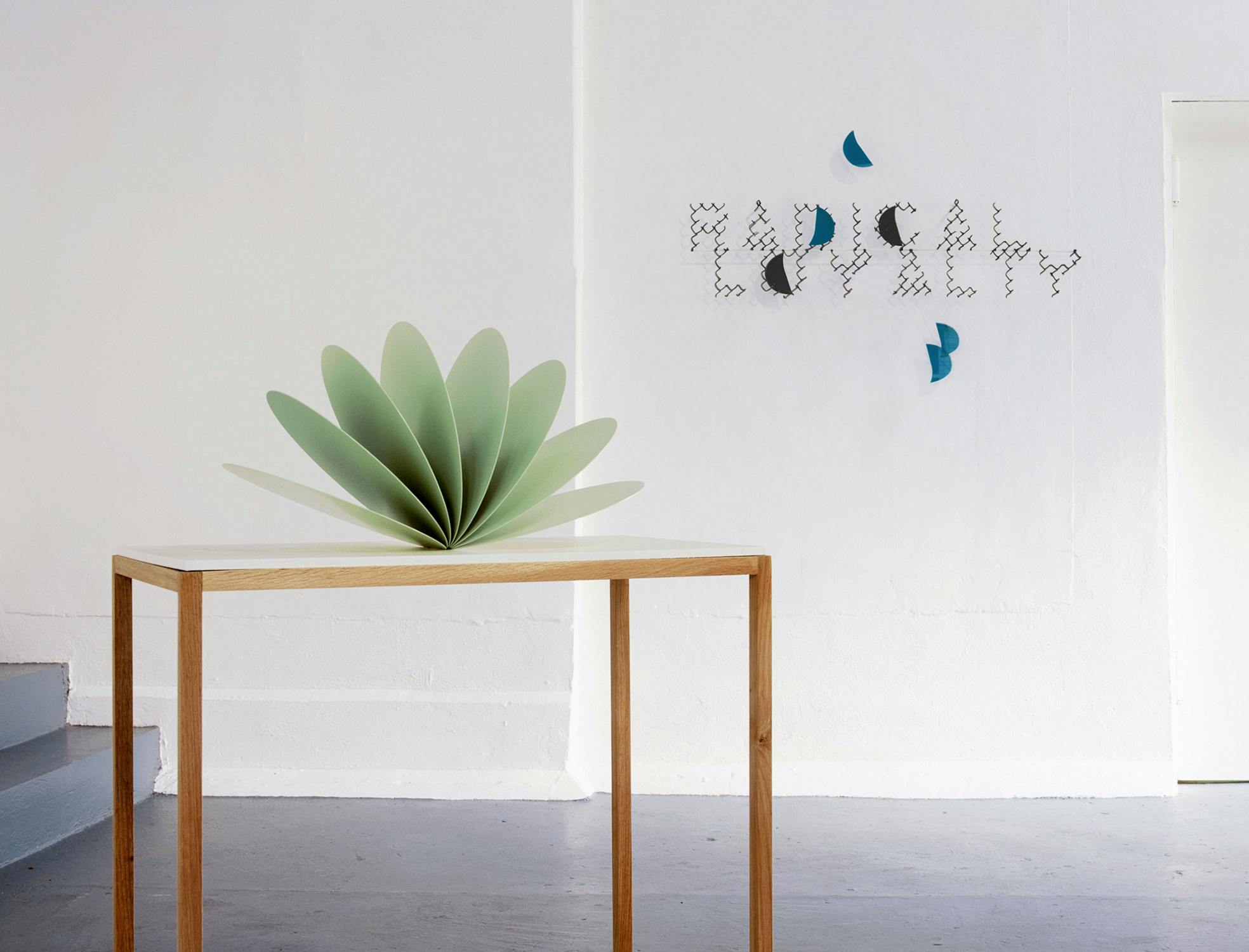 A minimalistic room features a wooden table with a fan-shaped green sculpture. The white wall behind the table displays fragmented text, which seems to spell out "RADICALLY." The space is airy and minimalist with a light and modern aesthetic.