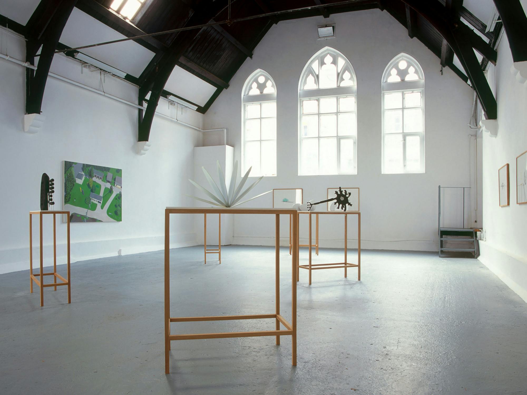 An art gallery with high ceilings and arched windows displays several sculptures on minimalist stands. The sculptures are varied in shape. A large painting hangs on the left wall, and framed artworks are arranged sparsely on the side walls. The space is well-lit.