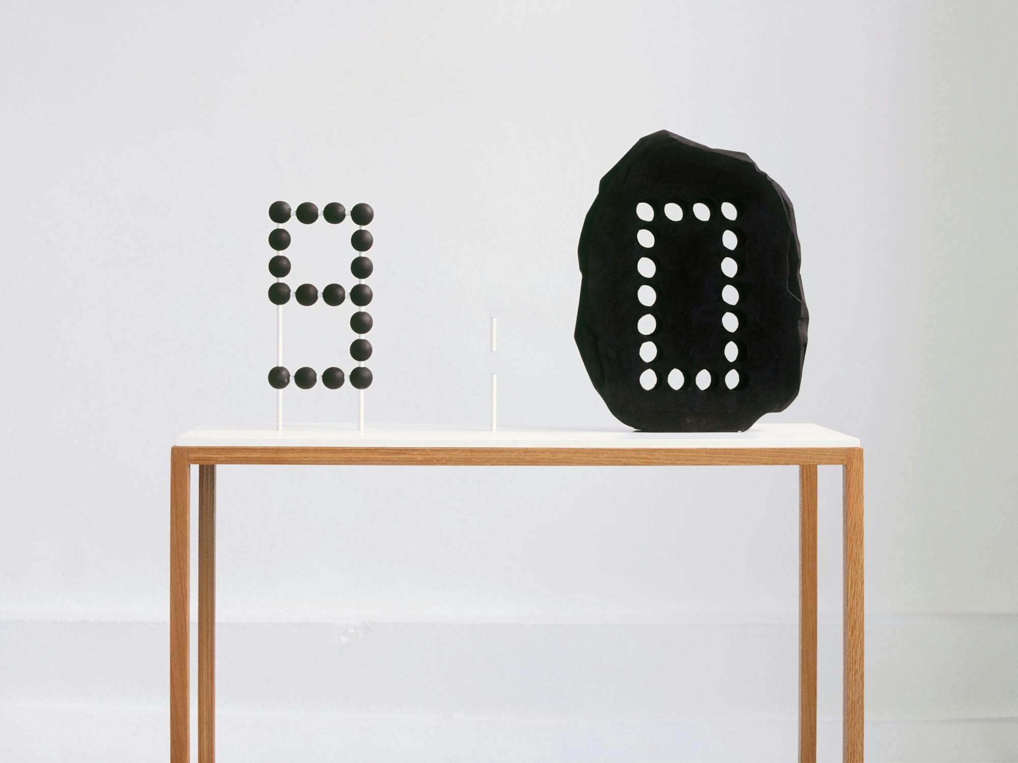 A minimalist setup shows two black sculptural pieces with white circular cutouts on a slim wooden table against a plain white background. One piece resembles the number "9" while the other resembles the number "0".