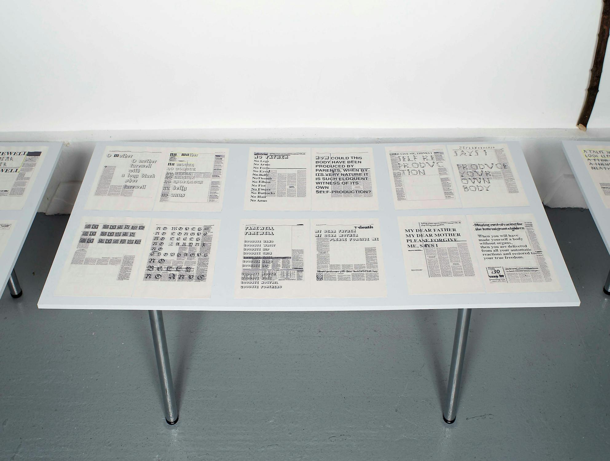 A display table showcasing a collection of vintage newspapers and documents. The texts and headlines are visible, though small and not fully readable. The table has a minimalistic design with sleek legs, positioned on a smooth gray floor against a plain white wall.