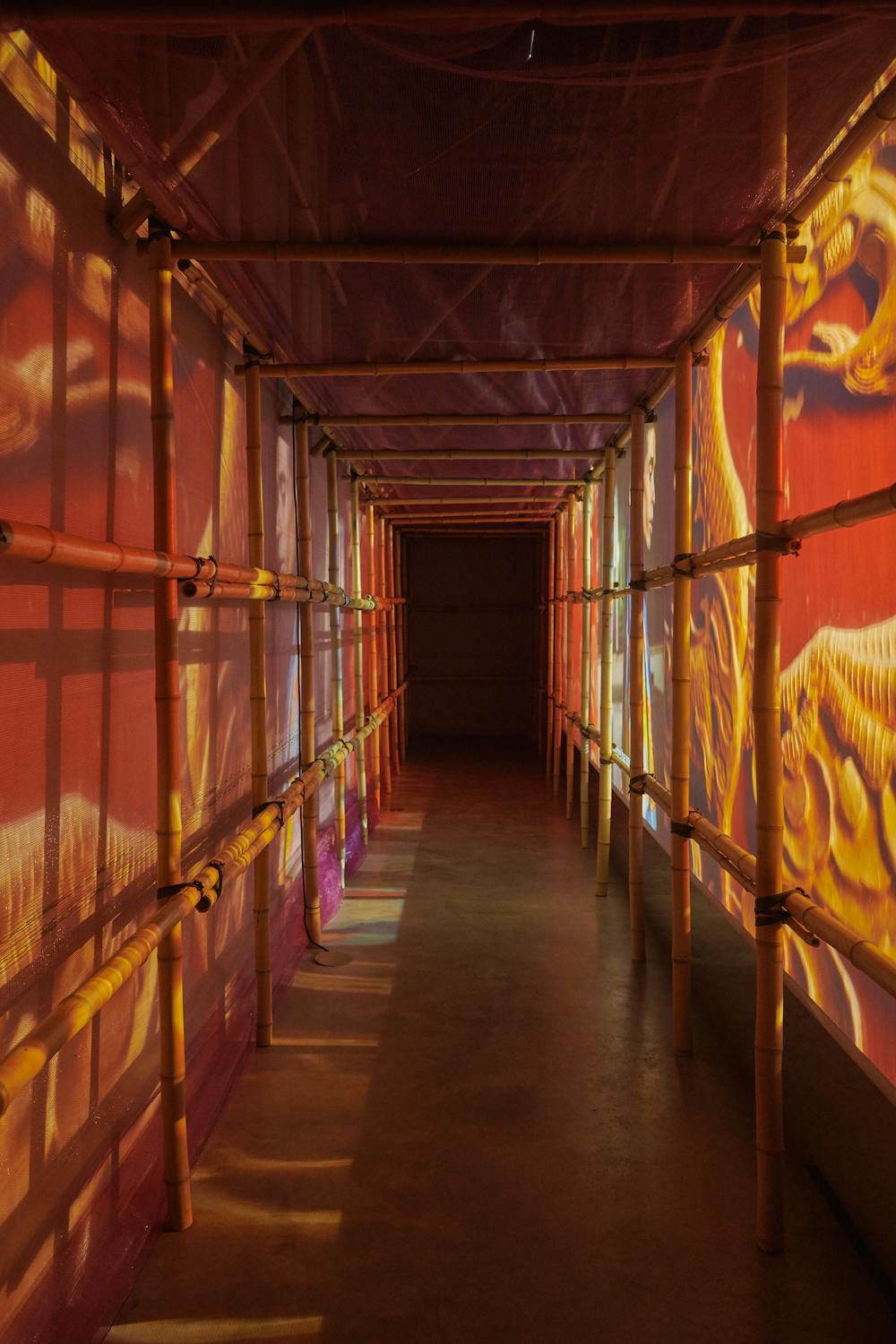 A narrow corridor made of bamboo poles is bathed in vibrant red and orange light projections, creating an ambient atmosphere. The light casts intricate patterns on the walls, adding to the visual richness of the space.