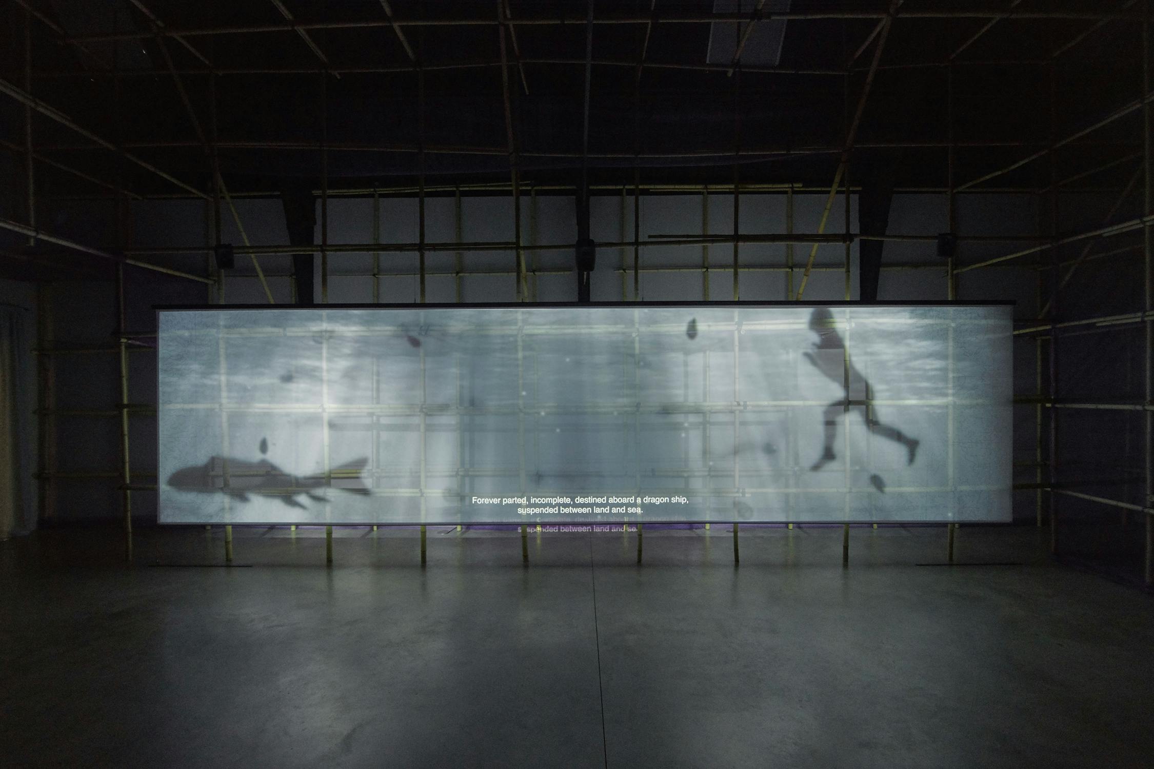 A large, illuminated screen displays an underwater scene in a dimly-lit gallery. Silhouettes of a person swimming and a fish can be seen. Bamboo scaffolding surrounds the screen, and the floor is concrete. Text appears at the bottom of the screen.