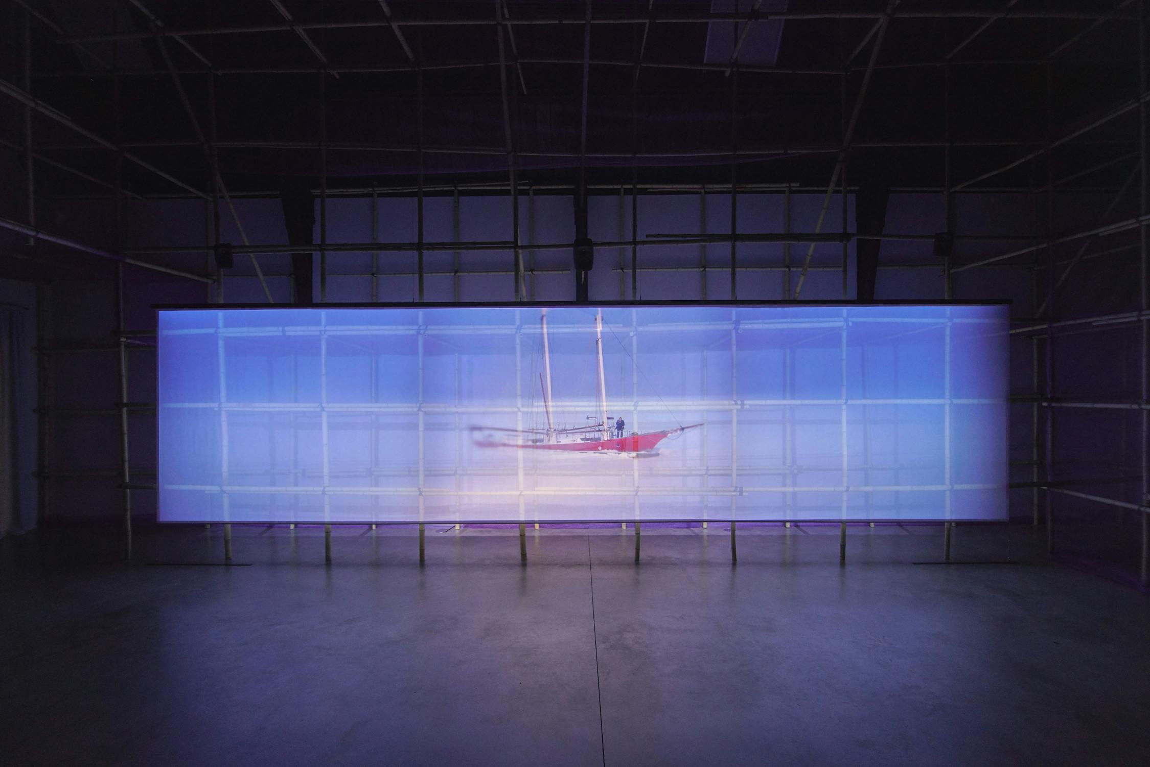 A digitally projected image of a red sailboat in a frozen sea against a large, rectangular screen in a dimly lit room. The image features a red and white boat centered on a light blue background. The screen is framed by a bamboo scaffolding structure.