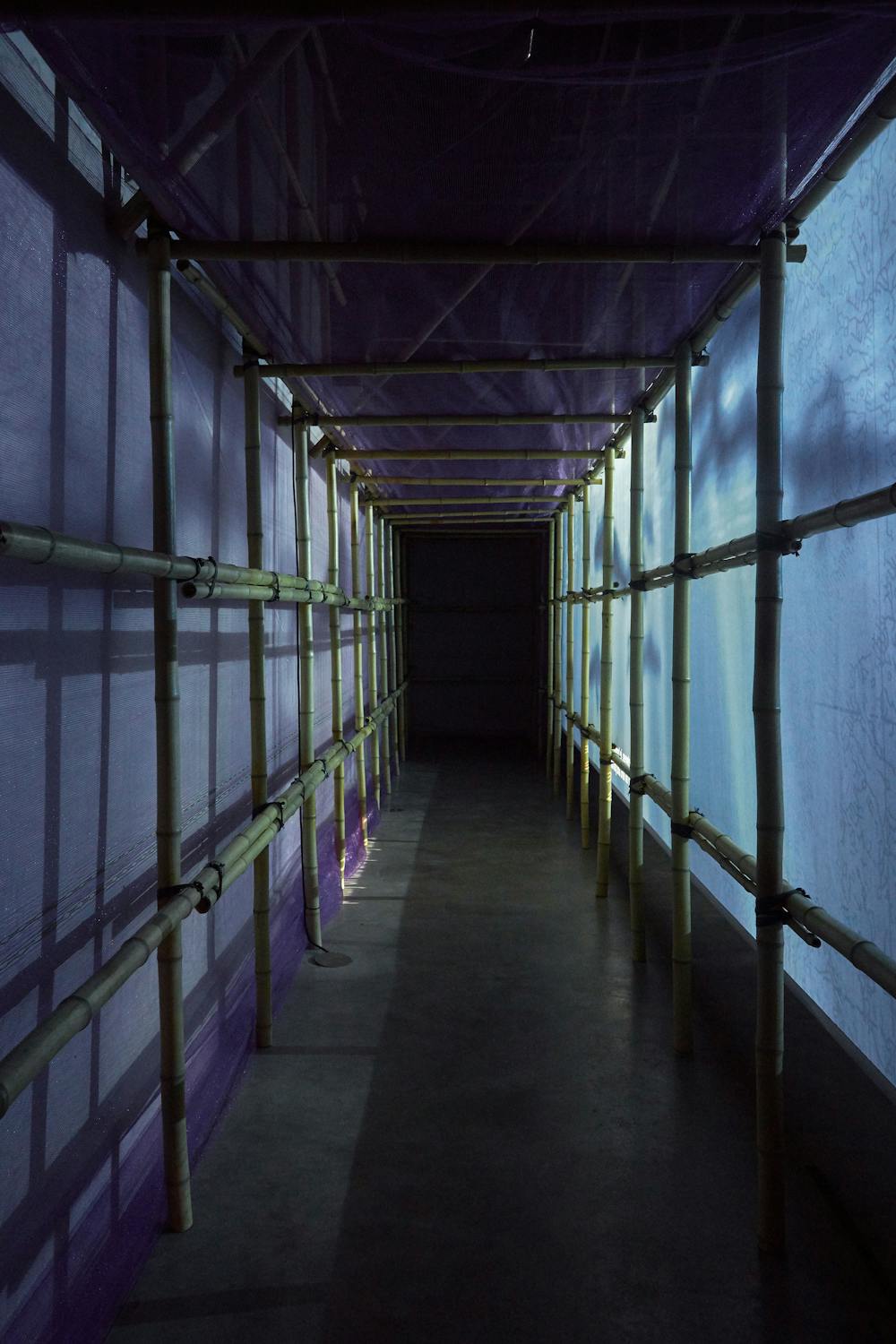 A narrow walkway constructed with bamboo poles and draped in purple mesh fabric creates a tunnel-like effect. The walkway is dimly lit, casting shadows and allowing light to softly filter through, leading to a darkened area at the end.