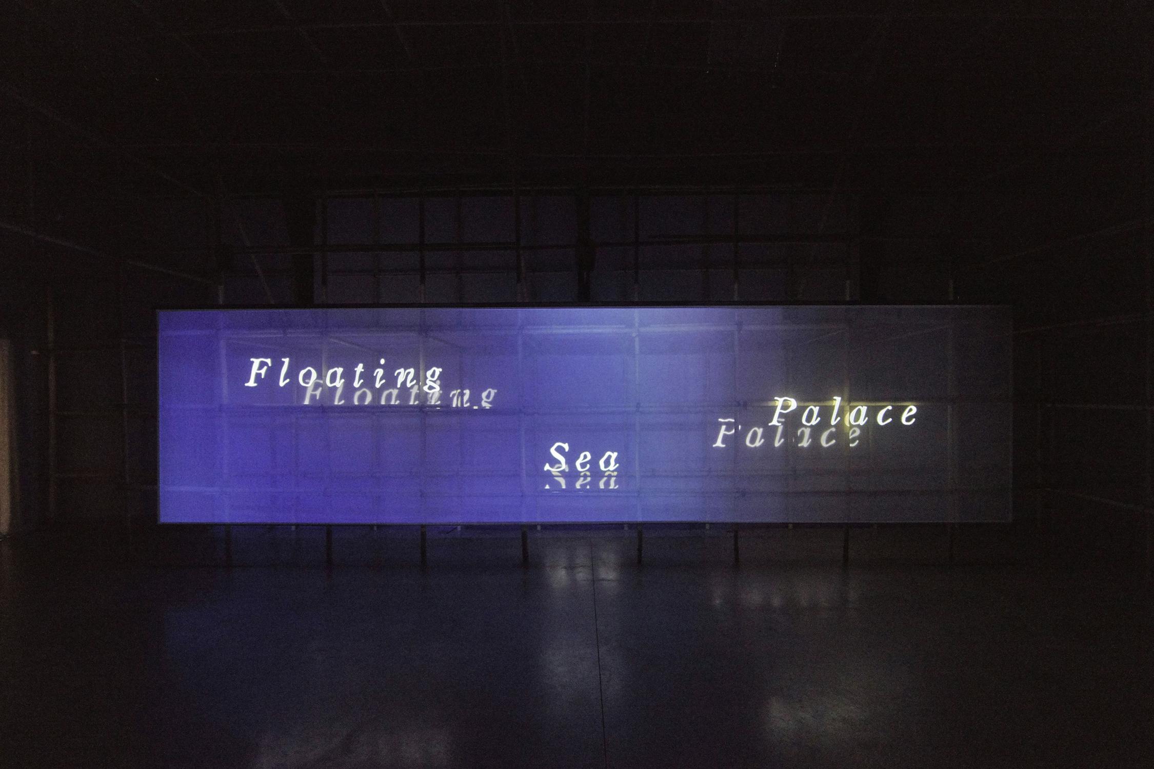 An art installation displays the words "Floating Sea Palace" on a large, semi-transparent screen. The text is distorted and illuminated against a dark background, creating an eerie and contemplative atmosphere.